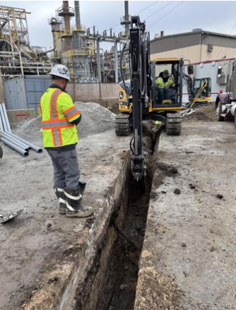 Construction workers on a work site with one using an excavator and another watching, with industrial structures in the background.