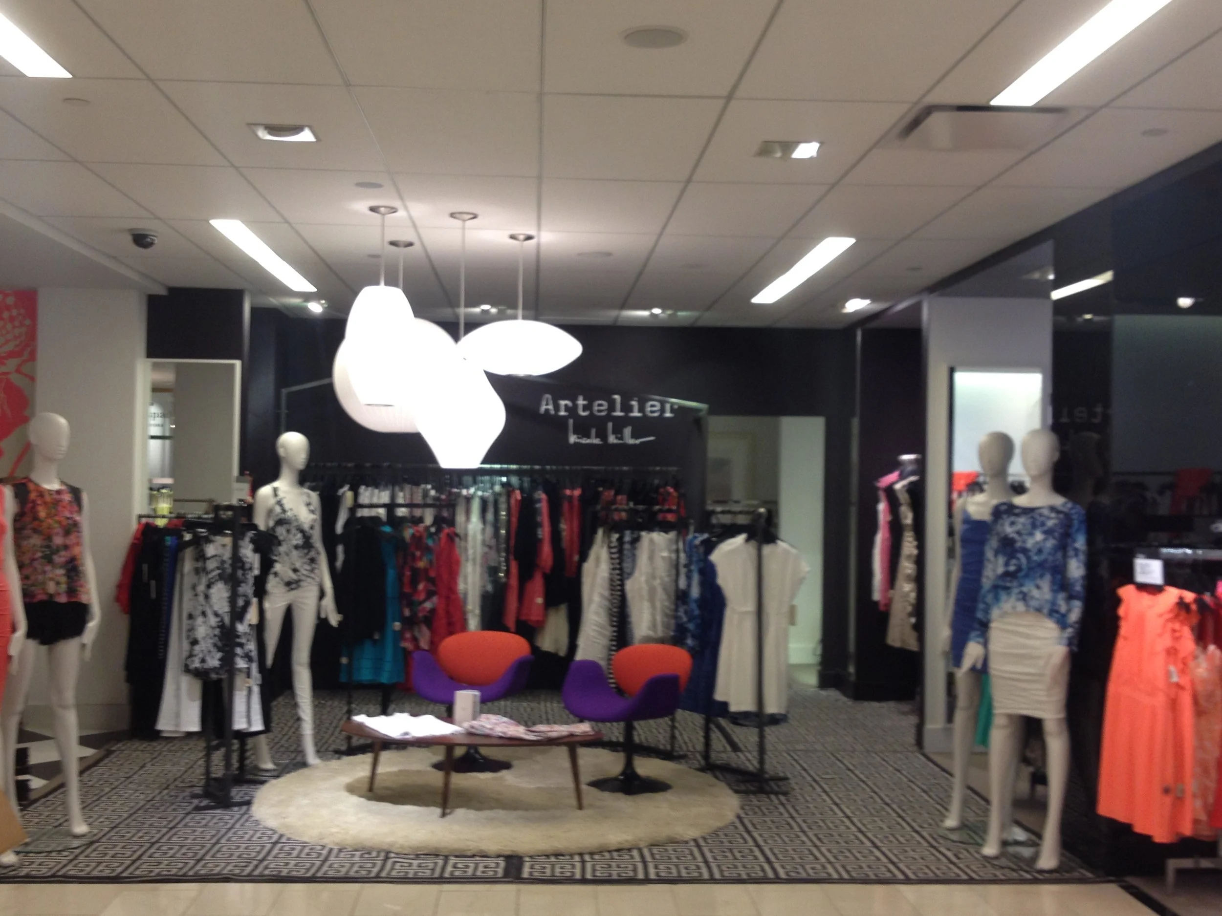 Mannequins dressed in colorful women's clothing in a boutique store with black walls, modern lighting, and a seating area with two orange chairs and a small table.