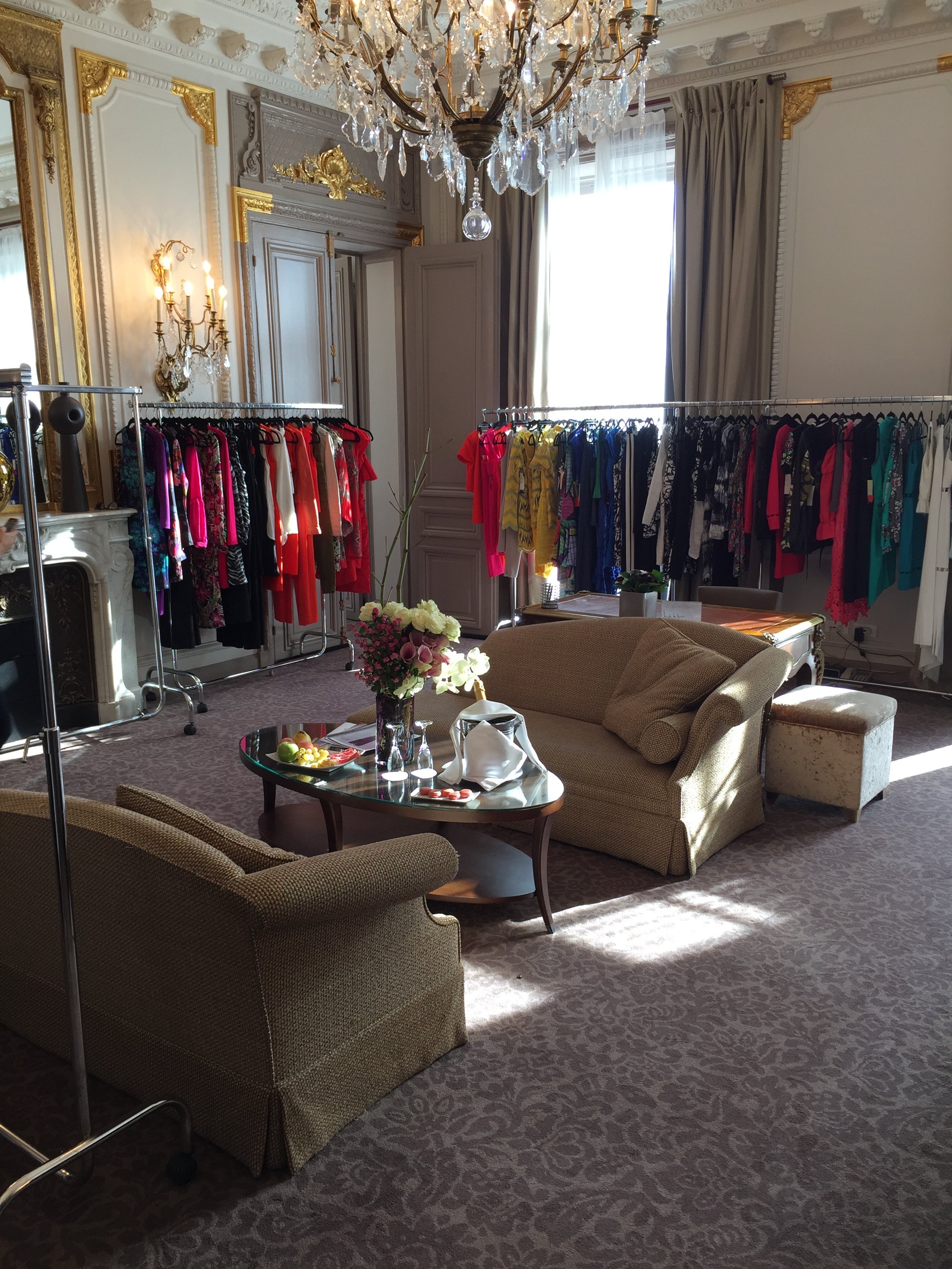 Elegant room with a chandelier, gold-accented walls, beige sofas, a coffee table with flowers and food, current clothing rack with colorful dresses, and sunlight coming through windows.
