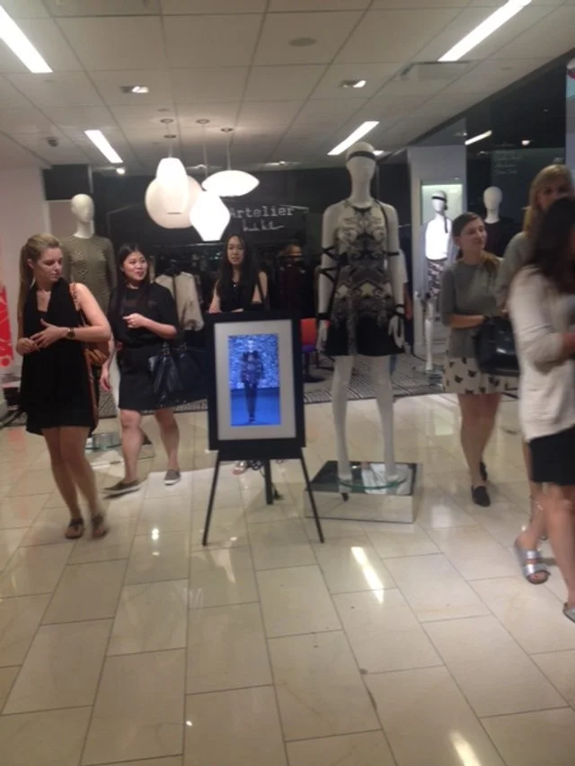 Mannequin dressed in black and beige patterned dress in shopping mall display with people walking by.