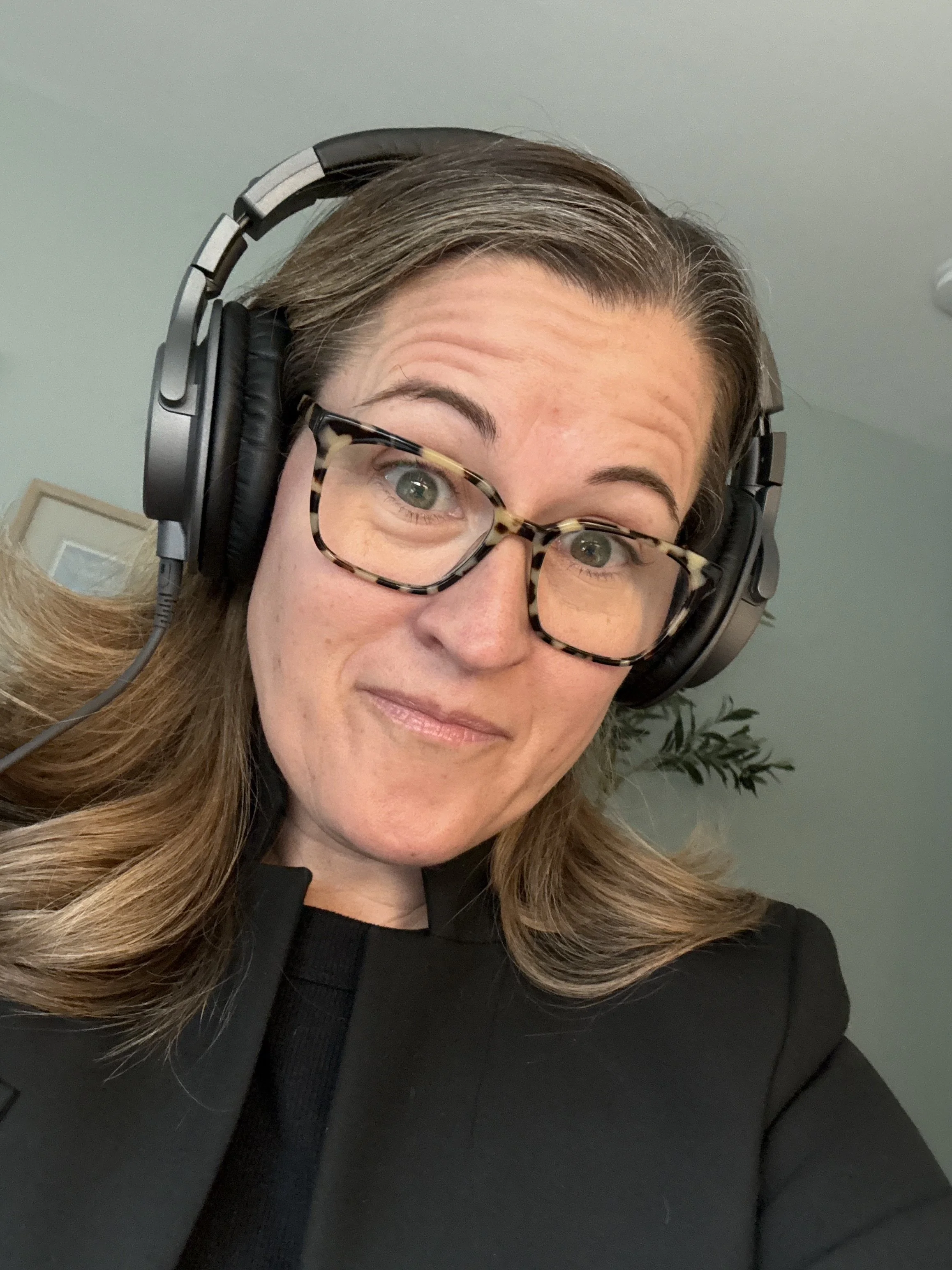A woman wearing glasses and large headphones, taking a selfie in a room with a light green wall and a plant in the background.