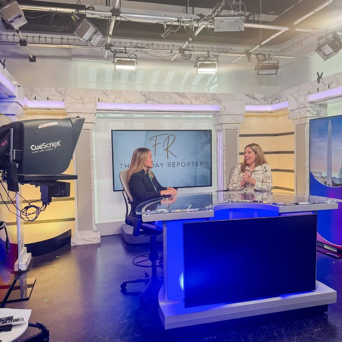 Two women seated at a news studio desk, smiling and engaging in conversation. Behind them, a large screen displays the text 'FR The Friday Reporter.' Studio lighting and equipment are visible overhead.