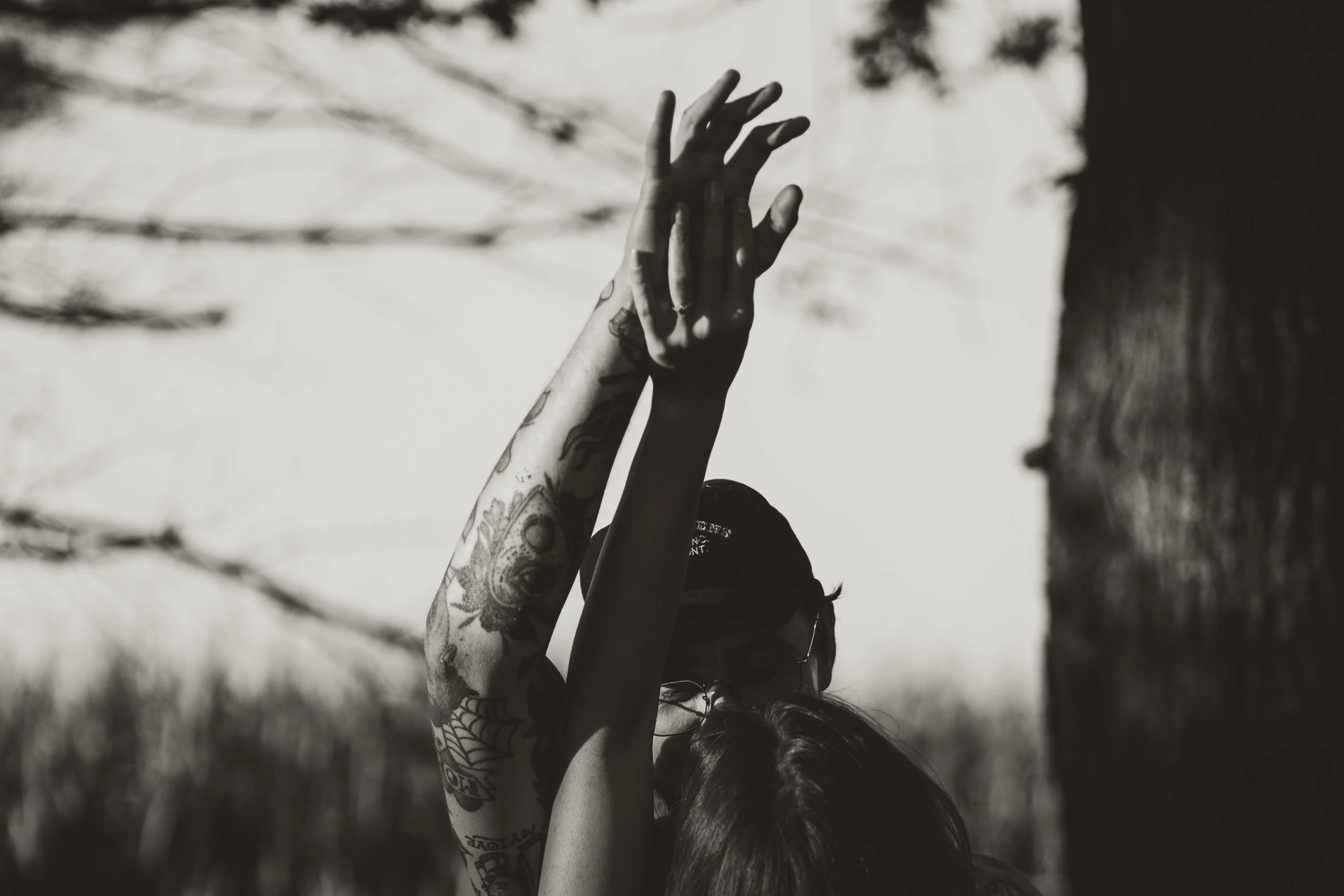 Two people with tattoos on their arms dancing outdoors near a tree, with one person wearing glasses and a baseball cap, raising their hands above their heads.