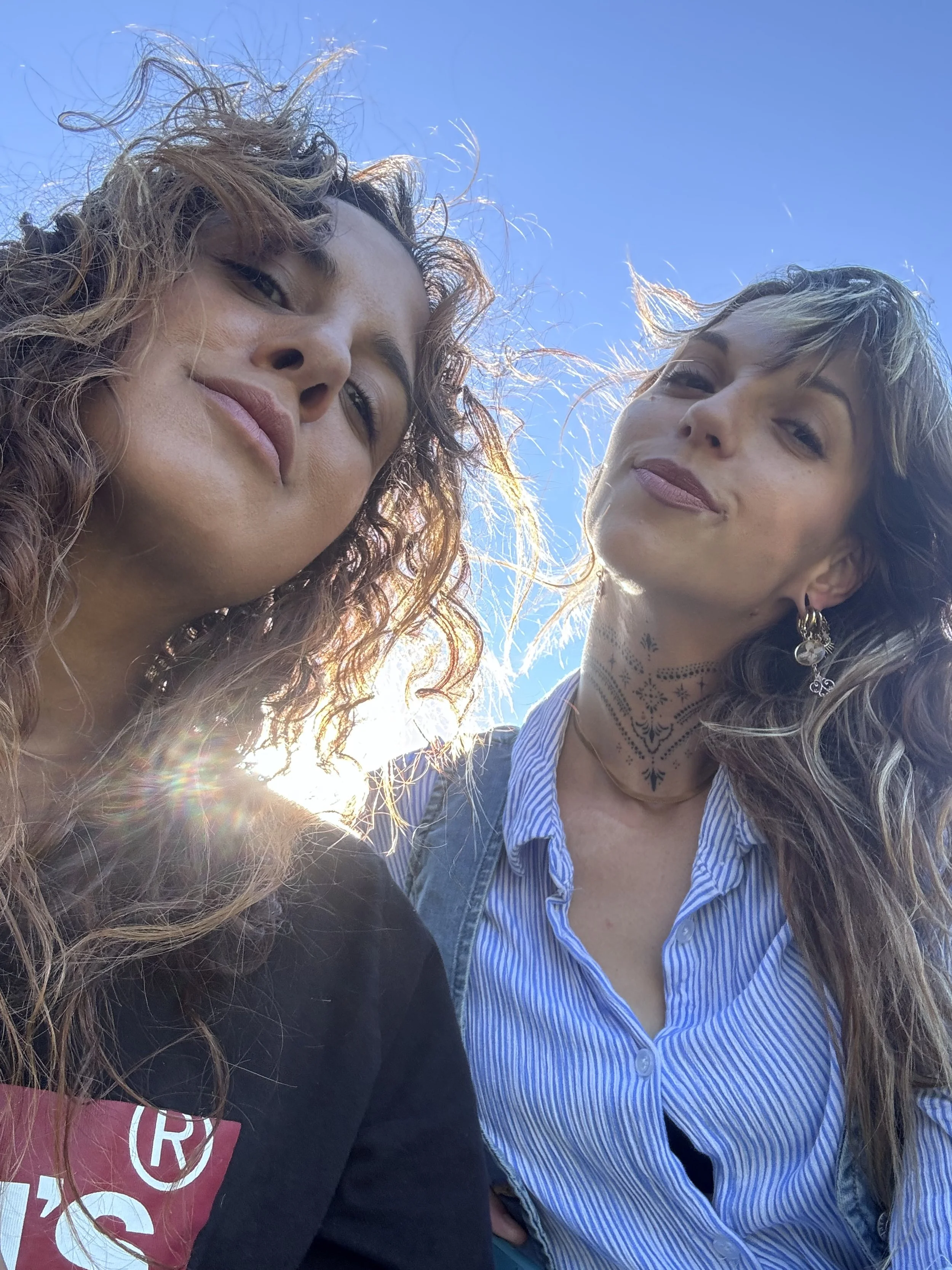 Two women taking a selfie outdoors under a clear blue sky, with sunlight behind them.