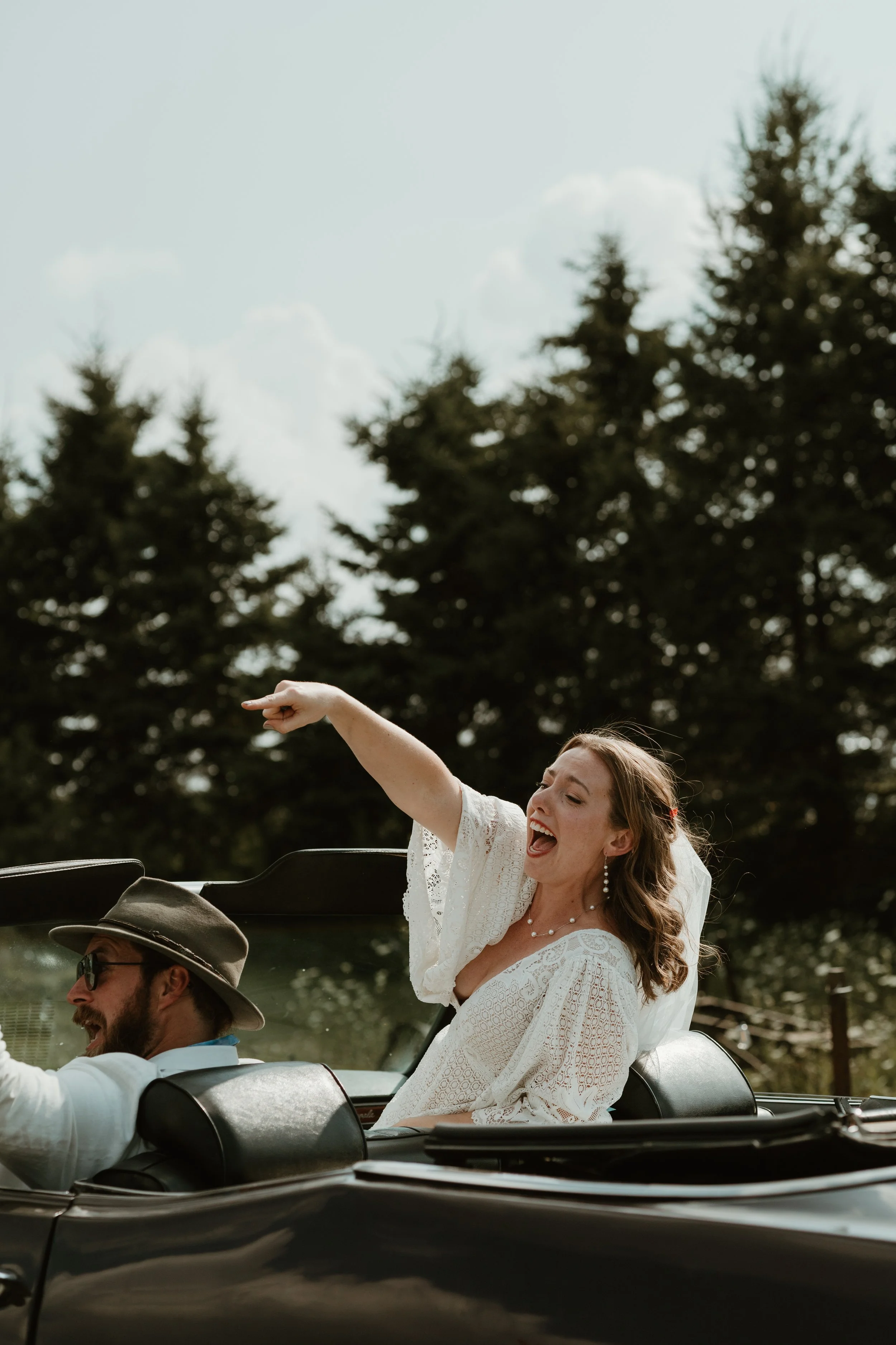 A woman in a white lace dress and pearl jewelry happily sitting in a black convertible car, pointing and laughing, with a man wearing sunglasses and a wide-brim hat driving the car, surrounded by tall trees under a partly cloudy sky.