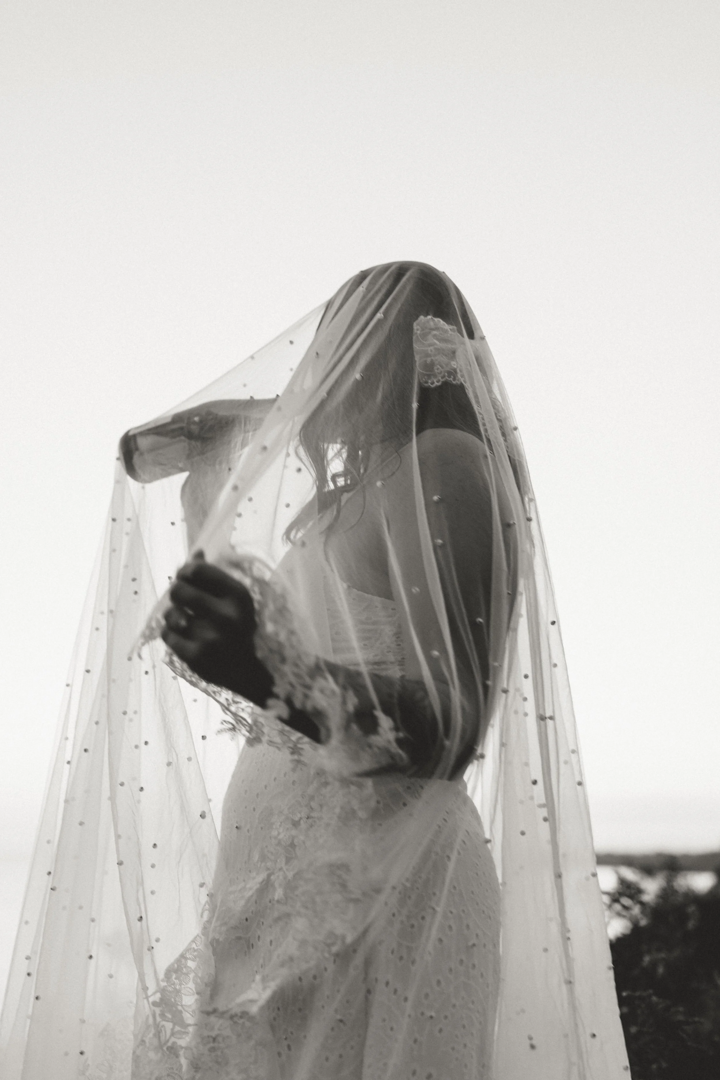 A woman in a lace dress is holding a sheer veil over her head, standing outdoors against a plain sky.