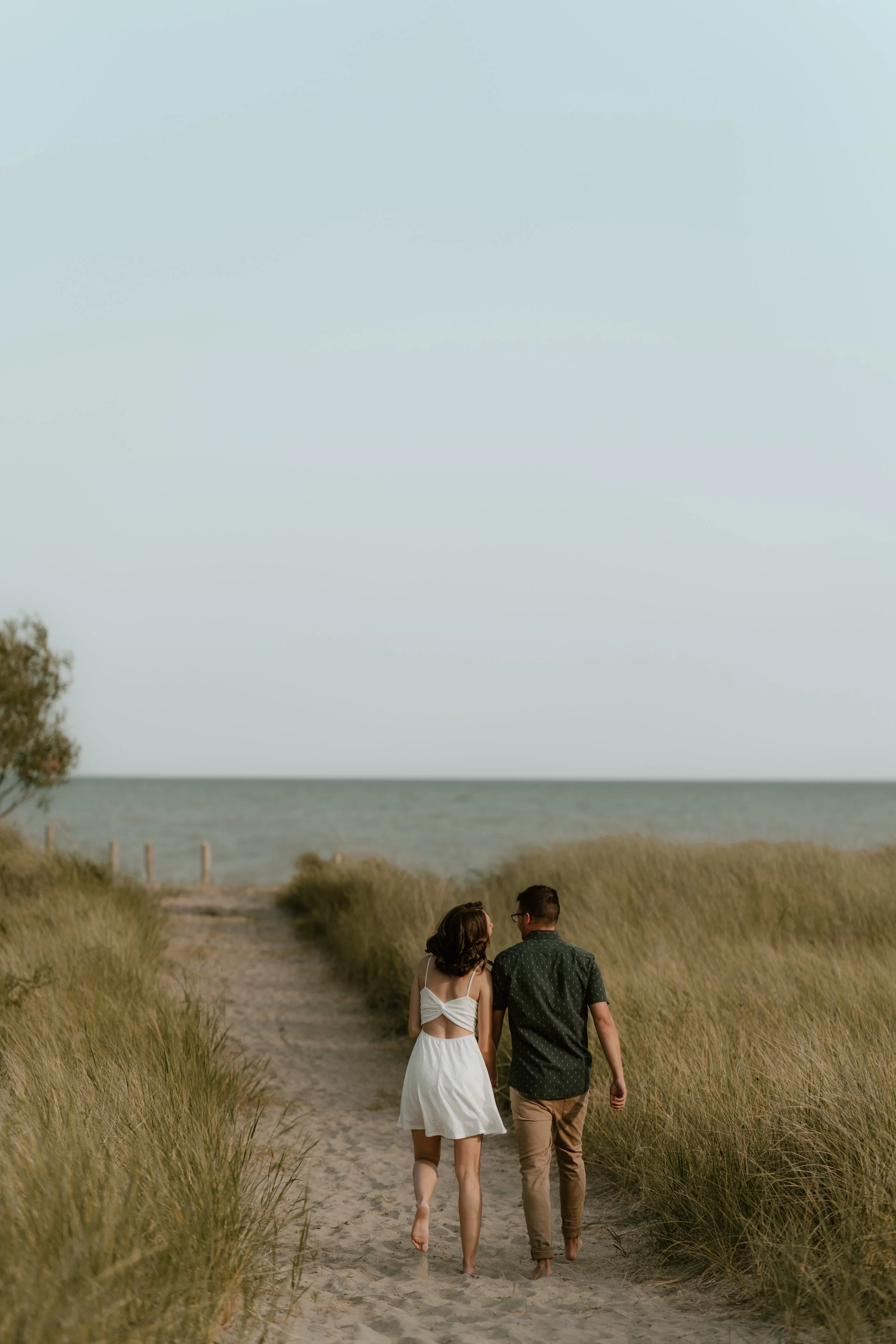 A young couple walking hand in hand on a sandy beach path lined with grass, with the ocean in the background on a clear day.