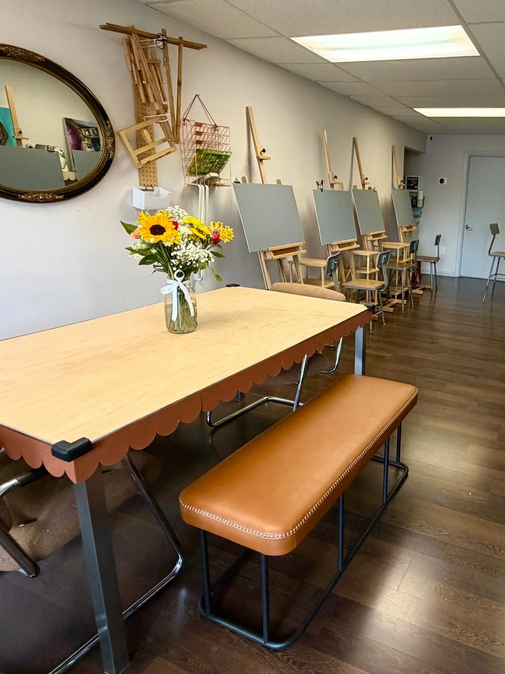 A wooden table with a vase of colorful flowers, including sunflowers and baby's breath, in the foreground. Behind the table are several artist's easels with blank canvases, positioned in a line across a room with a light-colored wall. There are chairs of various styles, including a brown leather bench, around the room, with a mirror and some wall-mounted storage visible in the background.
