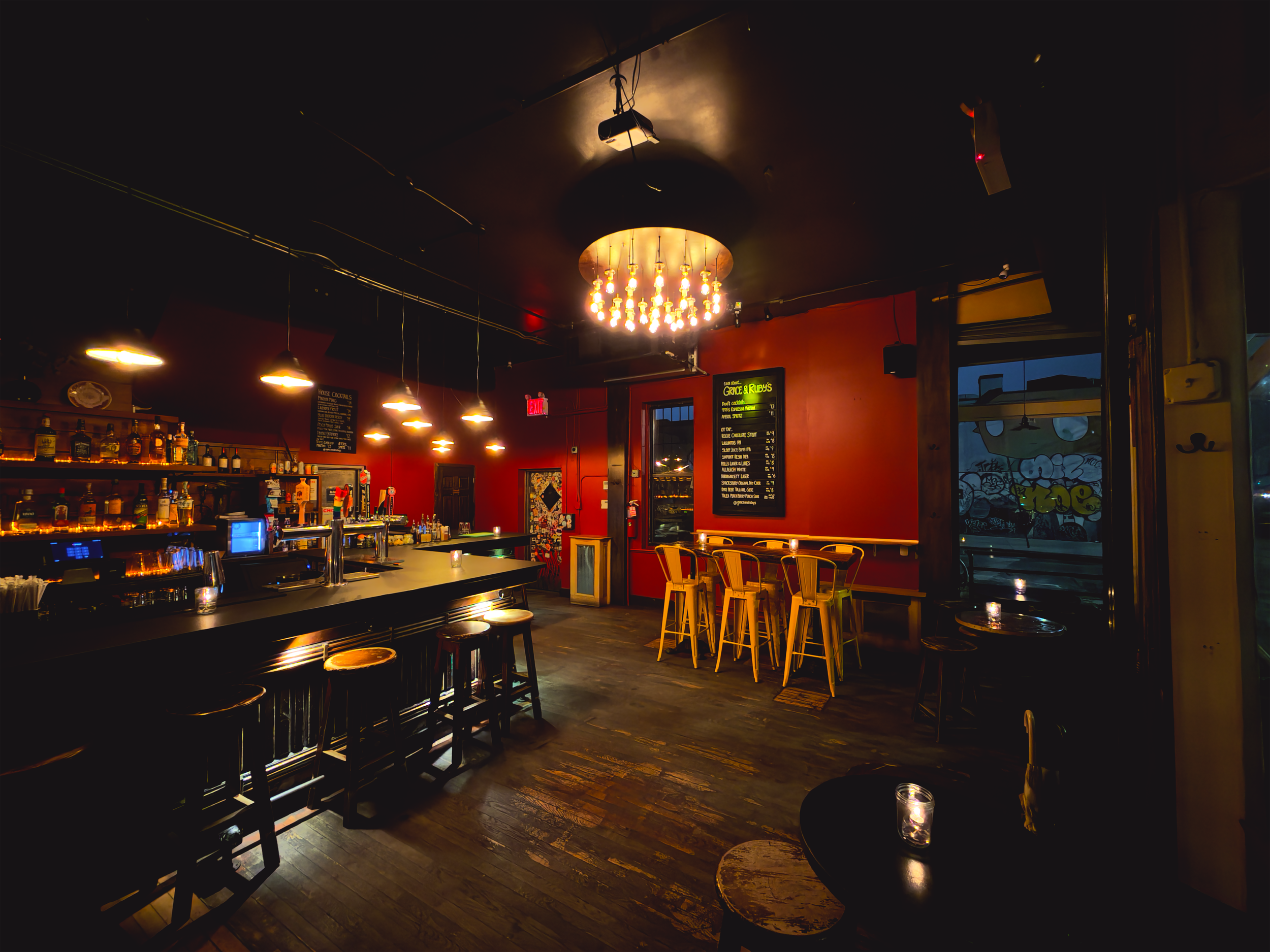 Empty bar with wooden stools, yellow chairs, a chalkboard menu, dim lighting, and graffiti on the window.