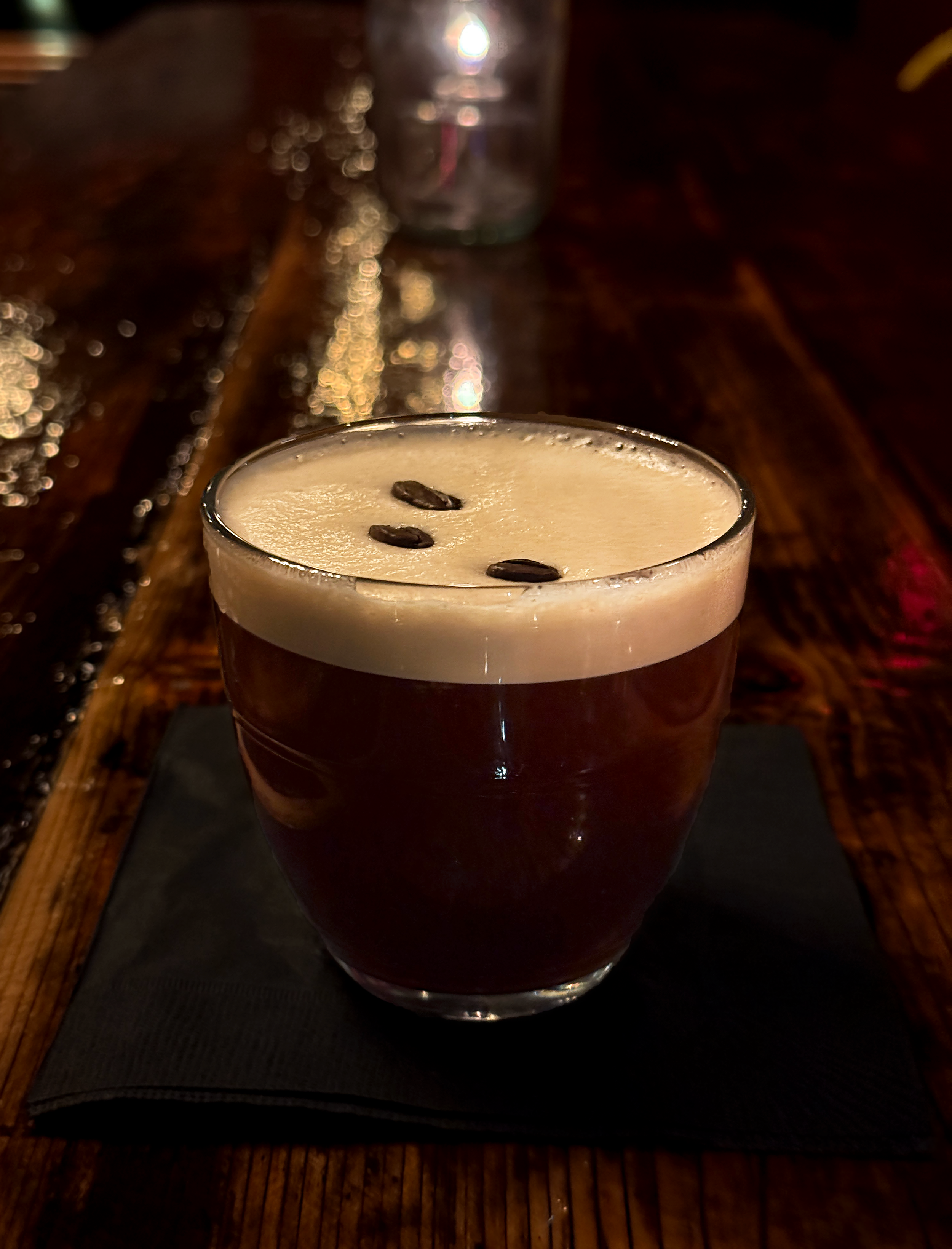 Espresso Martini, on a black napkin at a wooden bar or table.