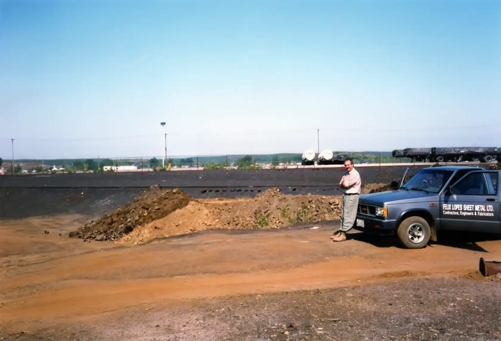 Man standing next to a pickup truck at a construction site with dirt piles, on a clear day.