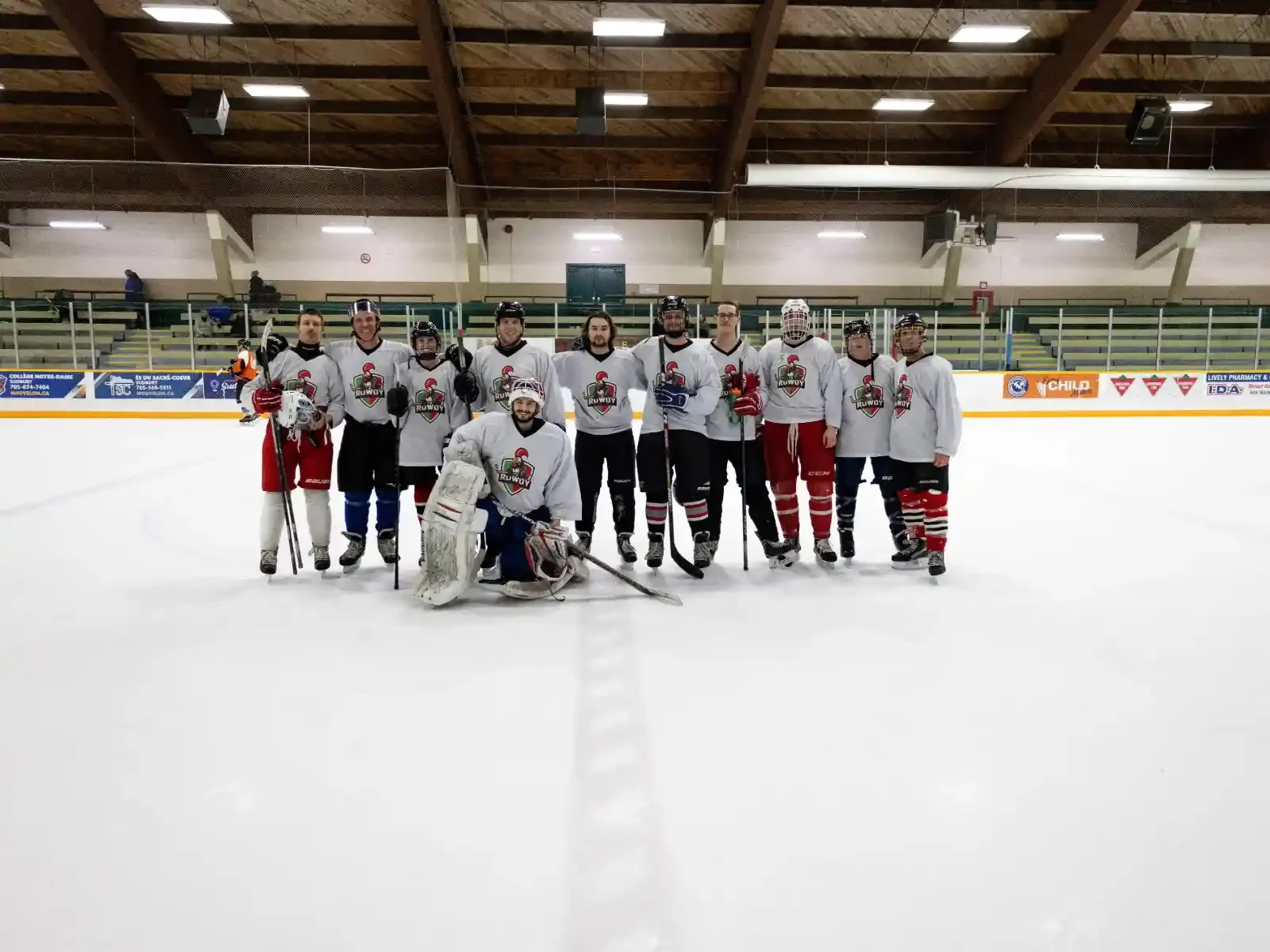 Hockey team posing on ice rink with players in uniform, some holding sticks, in an indoor ice hockey arena.