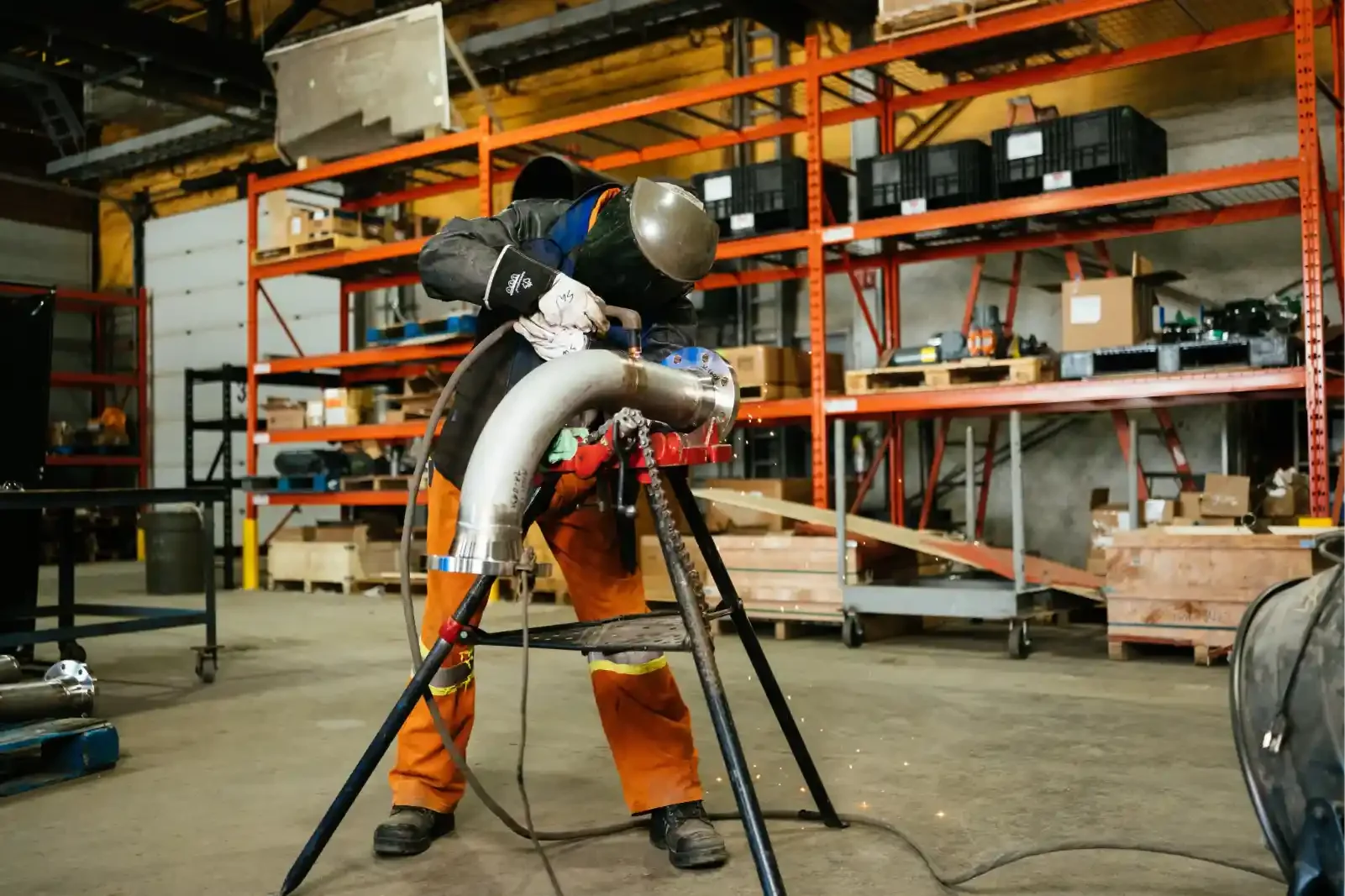 Worker welding a large metal pipe in an industrial workshop with orange shelving and various tools and boxes in the background.