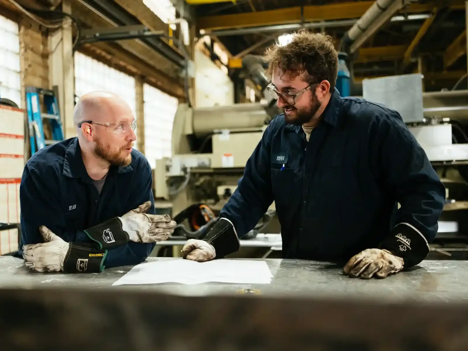 Two male workers in black uniforms and gloves at a workshop table, discussing papers.