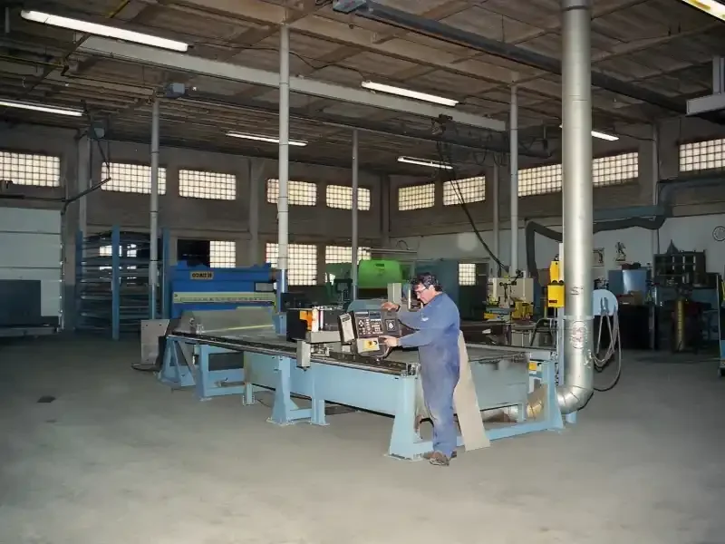A worker in blue overalls and safety glasses operating a CNC machine in an industrial workshop with high ceilings, large windows, and various manufacturing equipment.