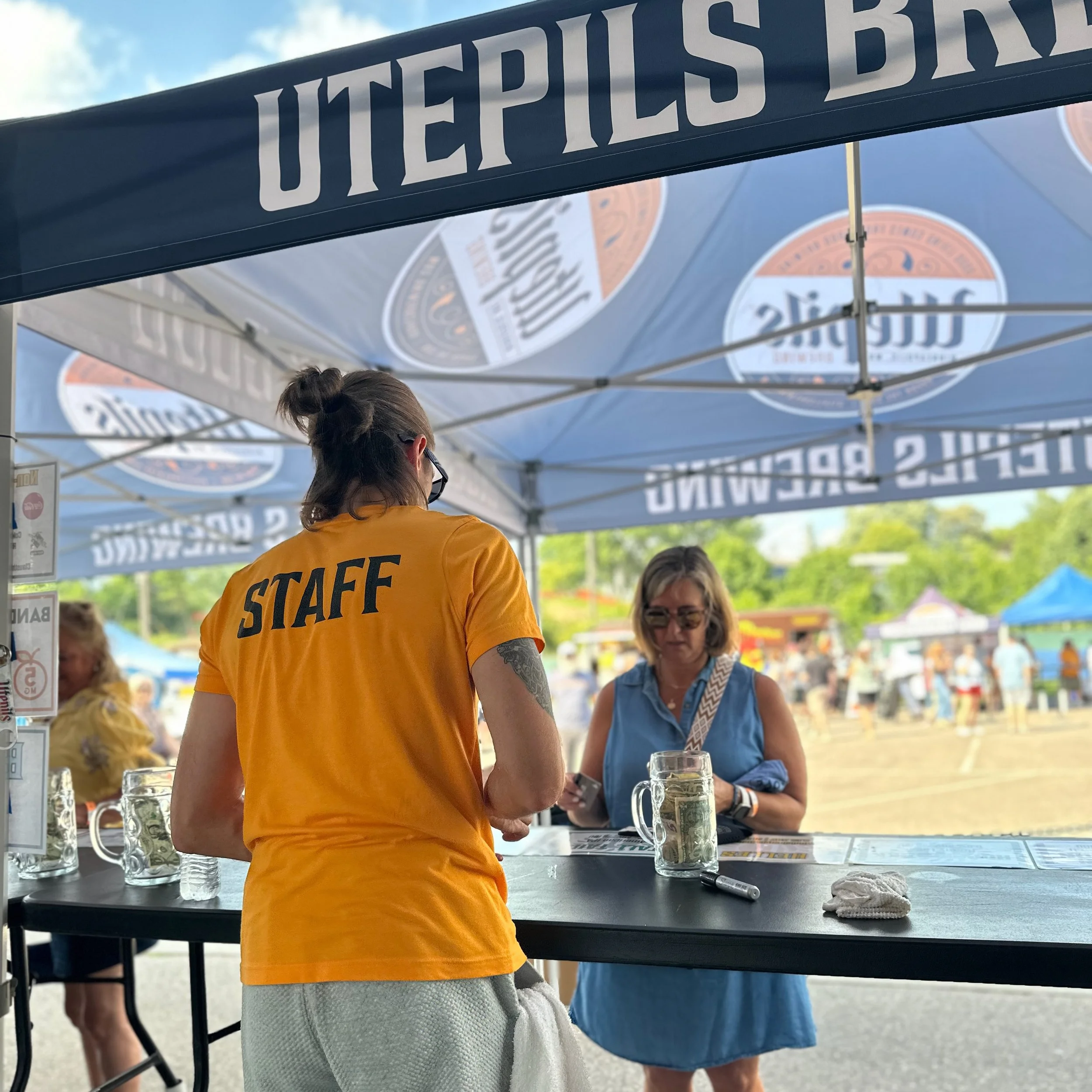 Staff member in a yellow shirt with 'STAFF' printed on the back assisting a woman at an outdoor event booth with a tent overhead.