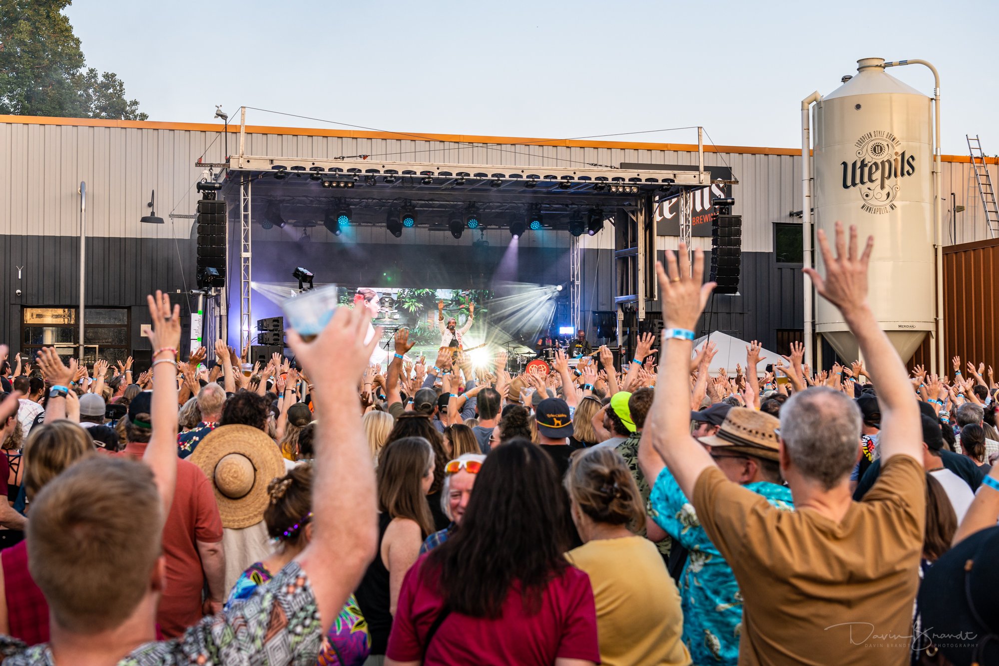 Crowd of people at an outdoor concert with hands raised, facing a stage with performers and bright lights.