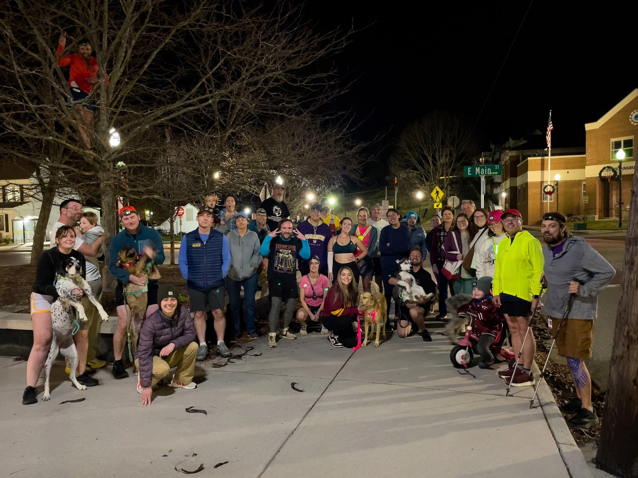 A large group of people and dogs gathered outdoors at night on a city street corner, with some members kneeling and others standing, some holding dogs and some raising their hands or making peace signs, under leafless trees and streetlights.