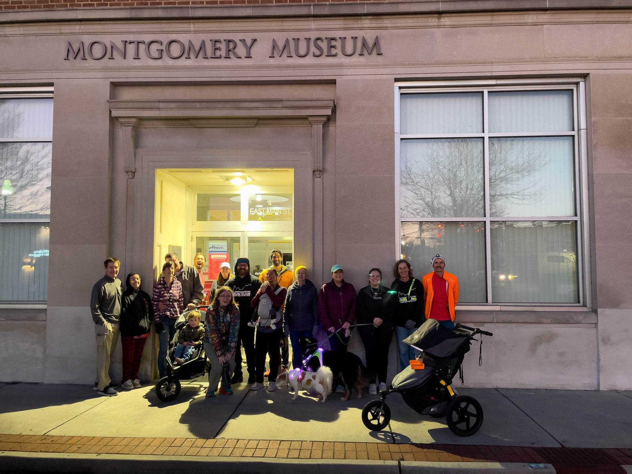 Group of people standing outside the Montgomery Museum at night, with some holding dogs and pushing a stroller, in front of the museum's entrance that is illuminated.