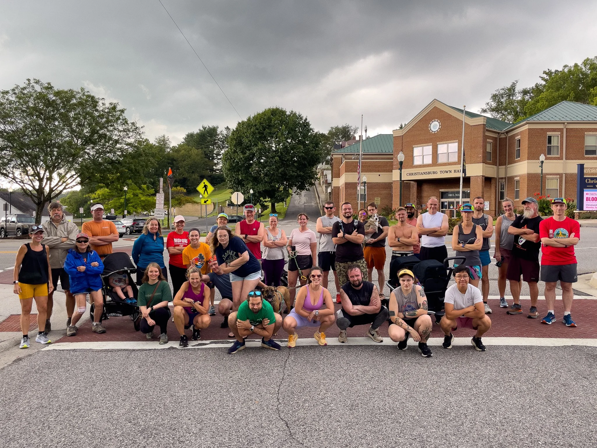 Group of people standing and crouching in front of Christansburg Town Hall, some with strollers and dogs, on a cloudy day.