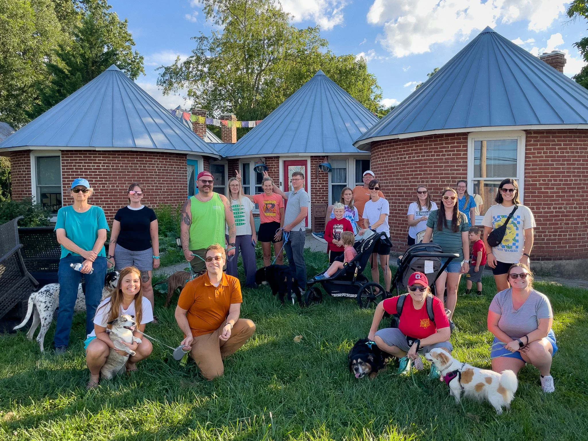 A group of people and dogs gathered outside of a brick house with three turreted roofs, some people are seated and some standing, with trees and a blue sky in the background.