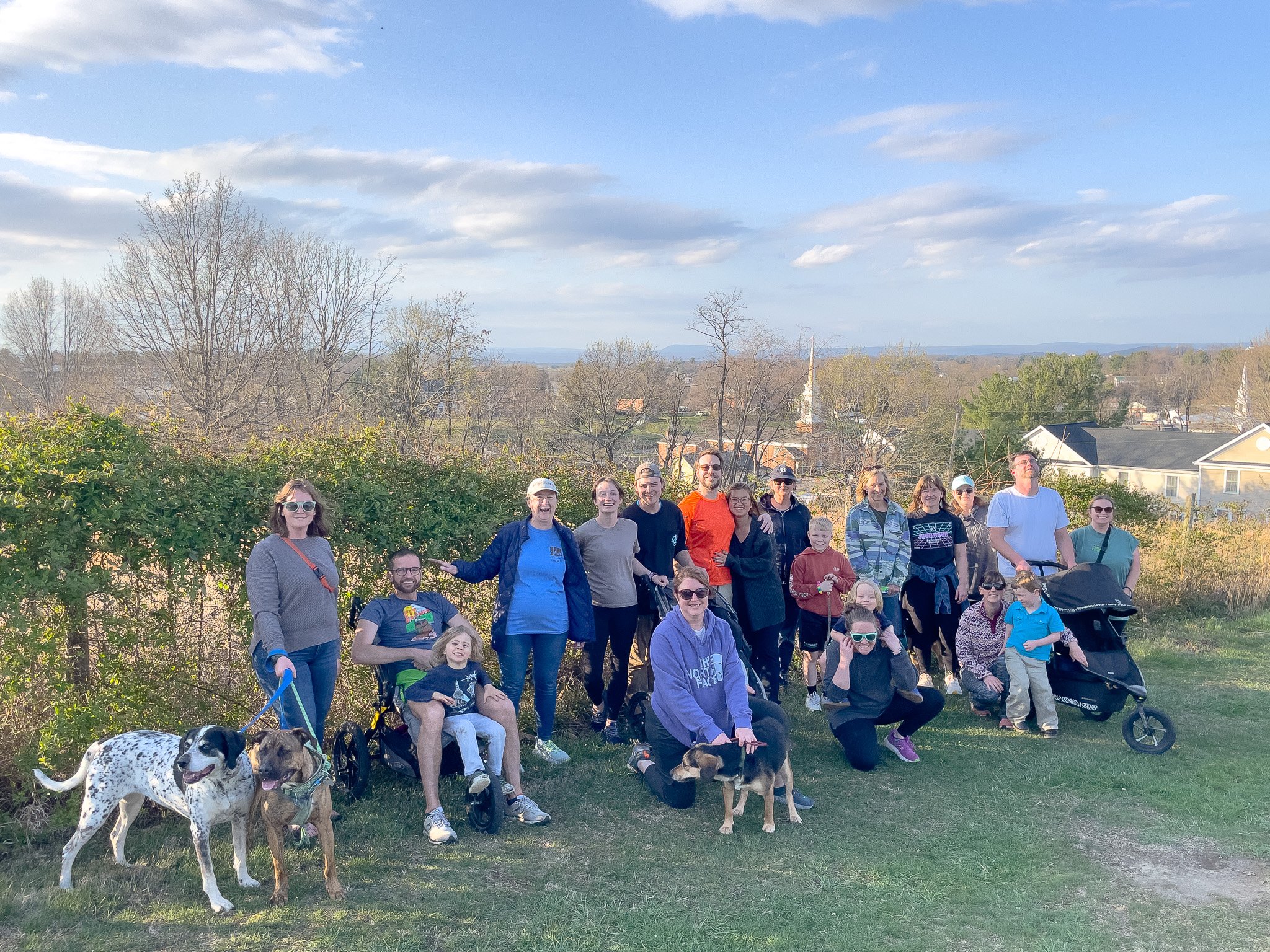 Group of people outdoors on a sunny day, some with dogs, standing on grass with trees and houses in the background.