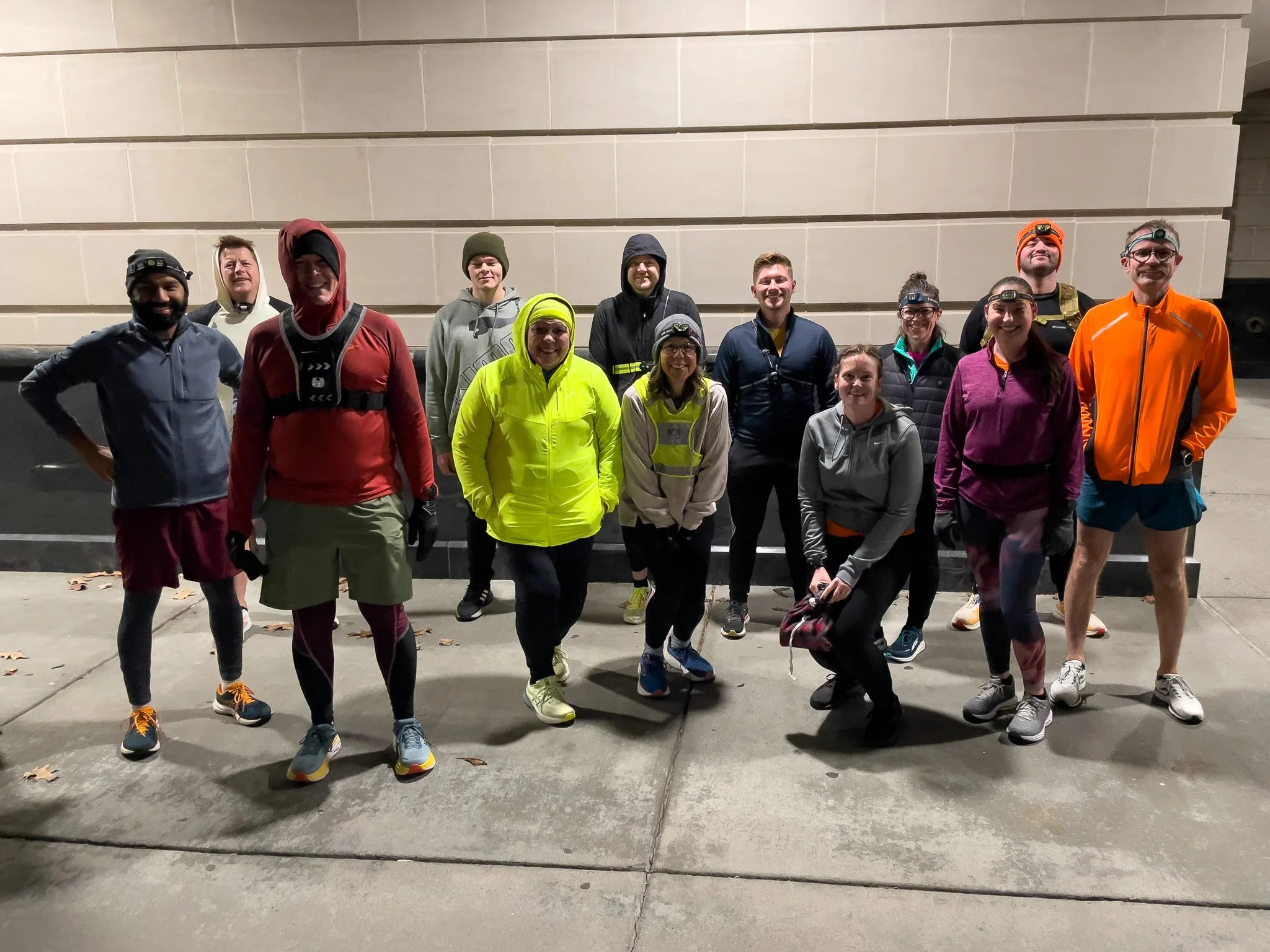 Group of fifteen people dressed in athletic or outdoor clothing, standing outdoors at night against a beige wall, some wearing headlamps, preparing for or finishing an outdoor activity or run.