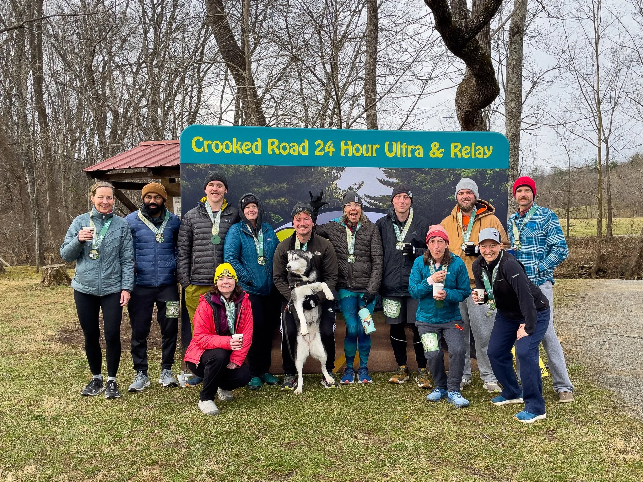 Group of people wearing medals standing outdoors in front of a large sign that reads 'Crooked Road 24 Hour Ultra & Relay.'