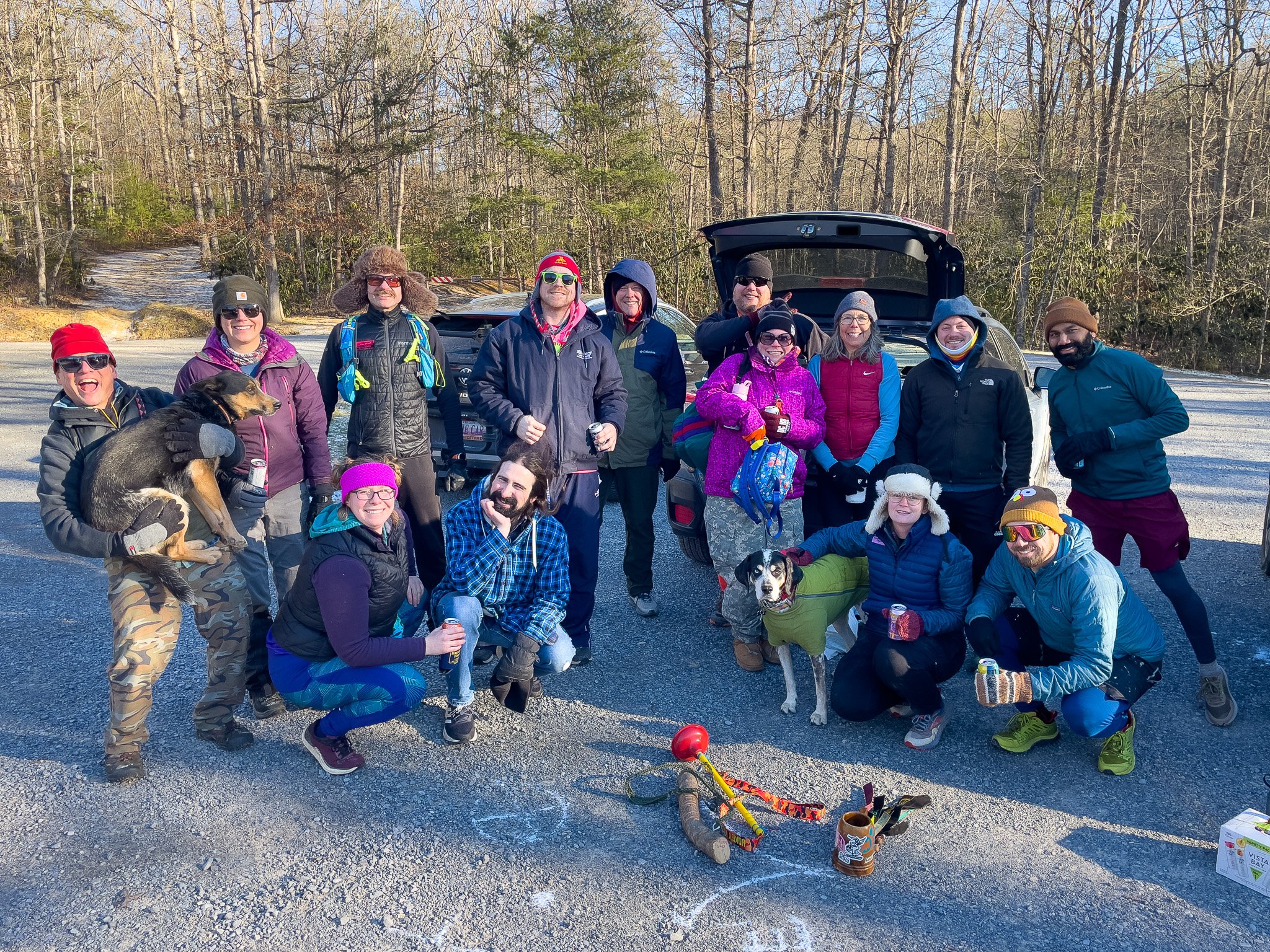Group of people and dogs outdoors on a sunny day, standing in front of cars in a wooded area, celebrating and smiling.