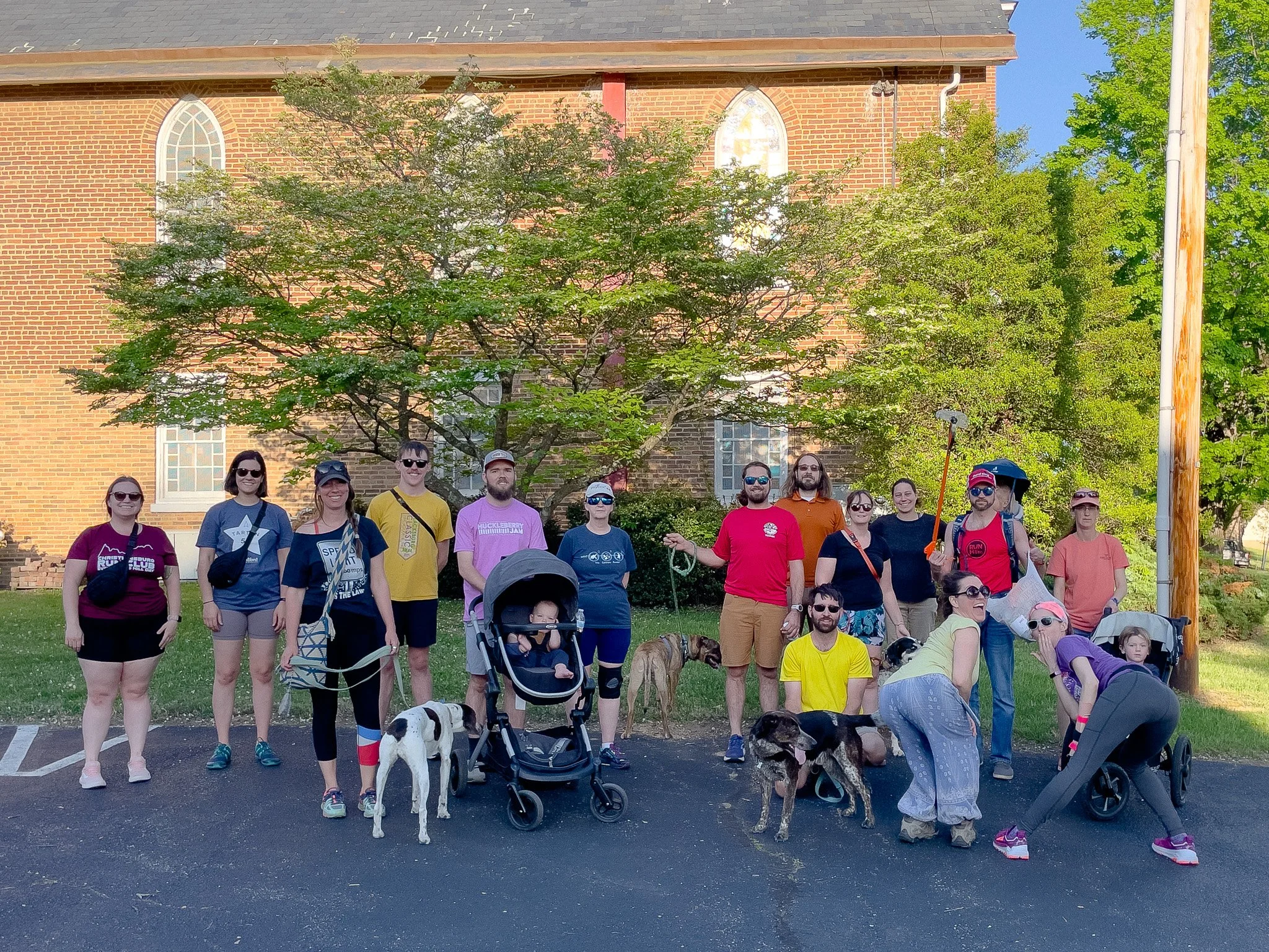 A group of people standing outdoors in front of a brick church building with large stained glass windows, along with several dogs, some on leashes. The group includes men and women of various ages, some wearing sunglasses, carrying backpacks, and pus