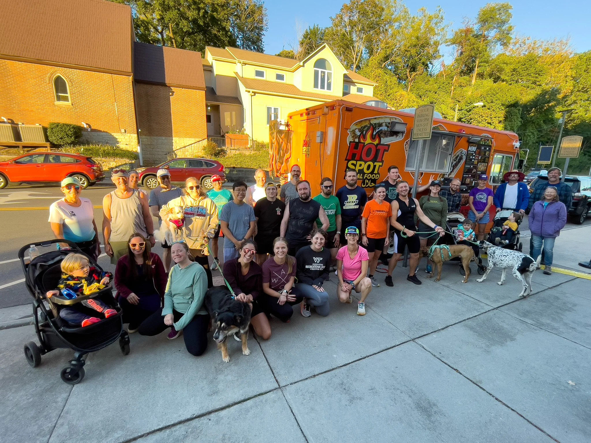 Group of people, including children and dogs, gathered outside in front of an orange food truck with 'HOT SPOT' written on it, during sunset with colorful lighting, in a neighborhood with houses and trees in the background.