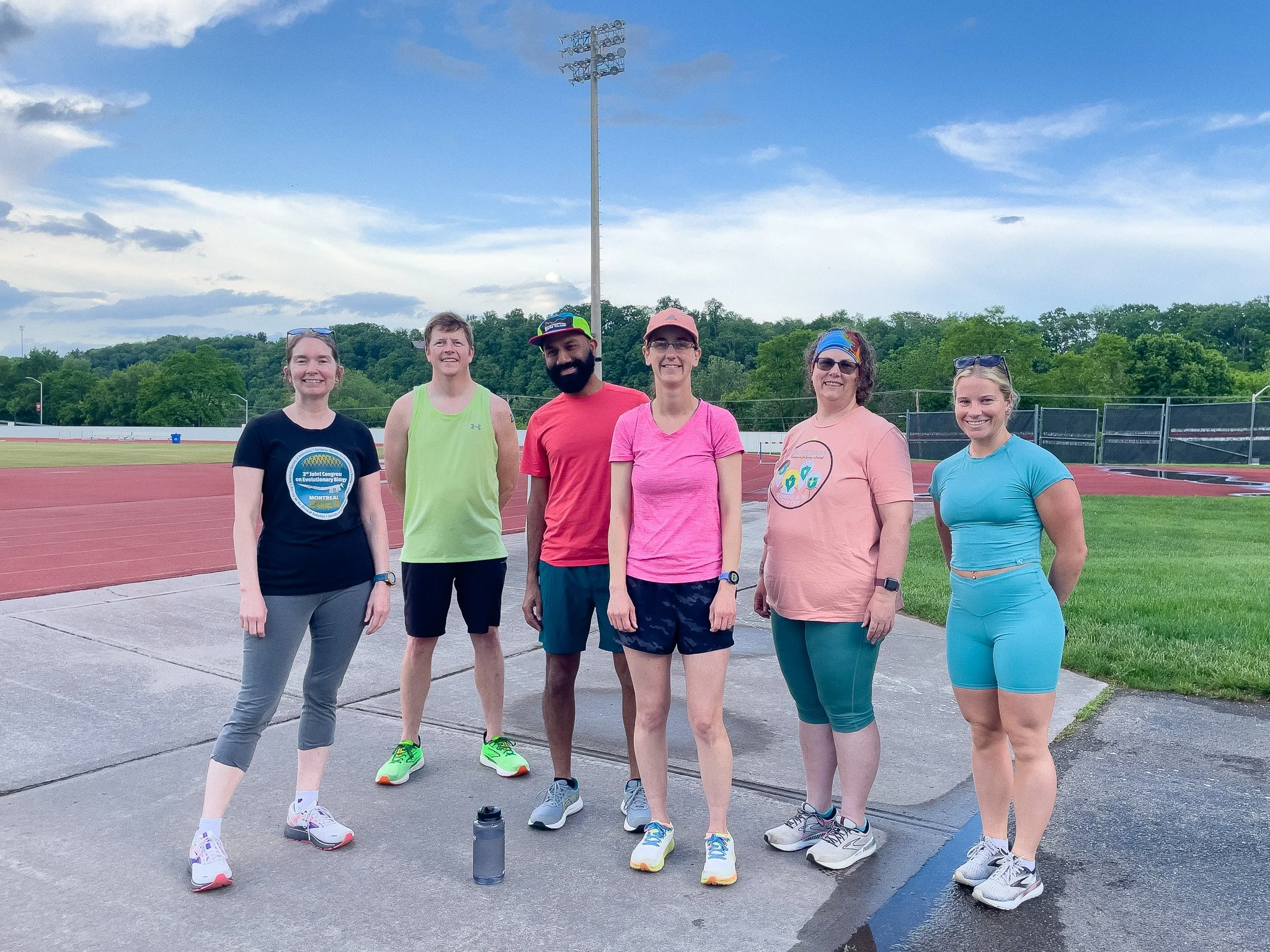 Six people standing on a track field with a running track, green grass, and trees in the background, smiling and looking at the camera.