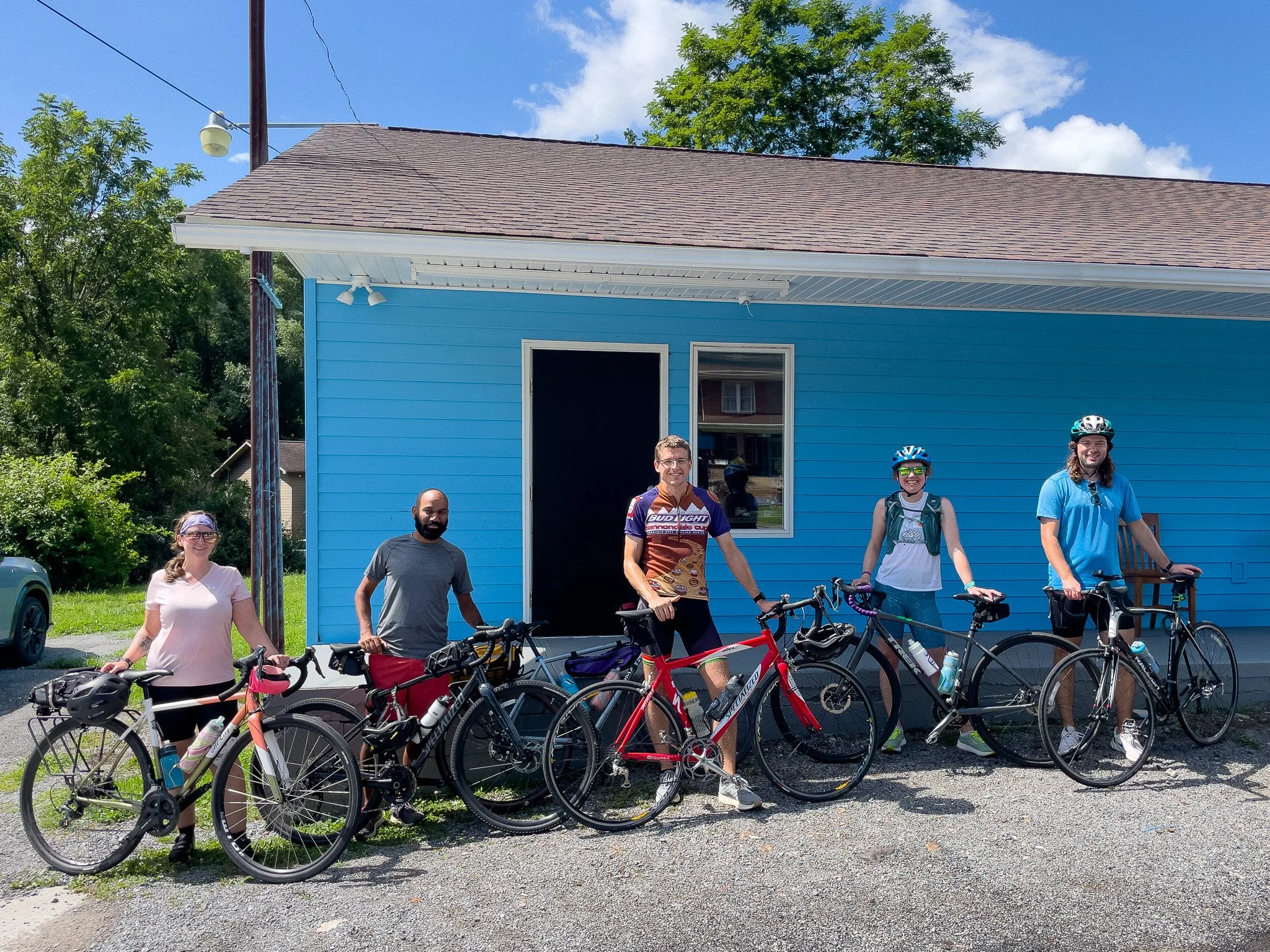 Five people standing with their bicycles in front of a blue building on a sunny day, wearing cycling gear and helmets.