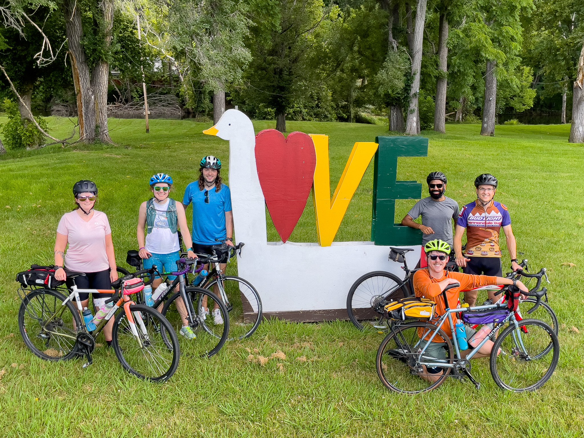 Group of six people wearing helmets with bicycles, standing in front of a large love sign with a duck head, a red heart, and green and yellow letters spelling 'LOVE'. The scene is set outdoors with grass and trees in the background.