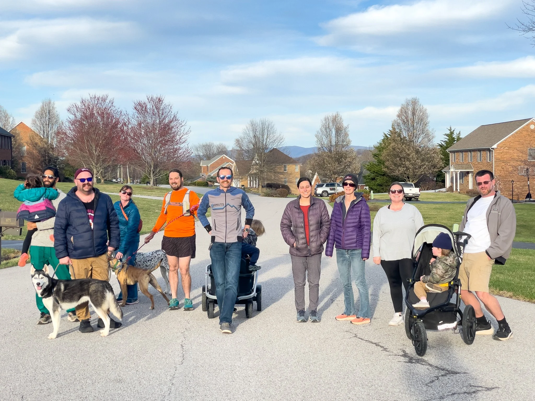 A group of people standing outdoors on a sunny day, with dogs and children, in a neighborhood with houses and trees in the background.
