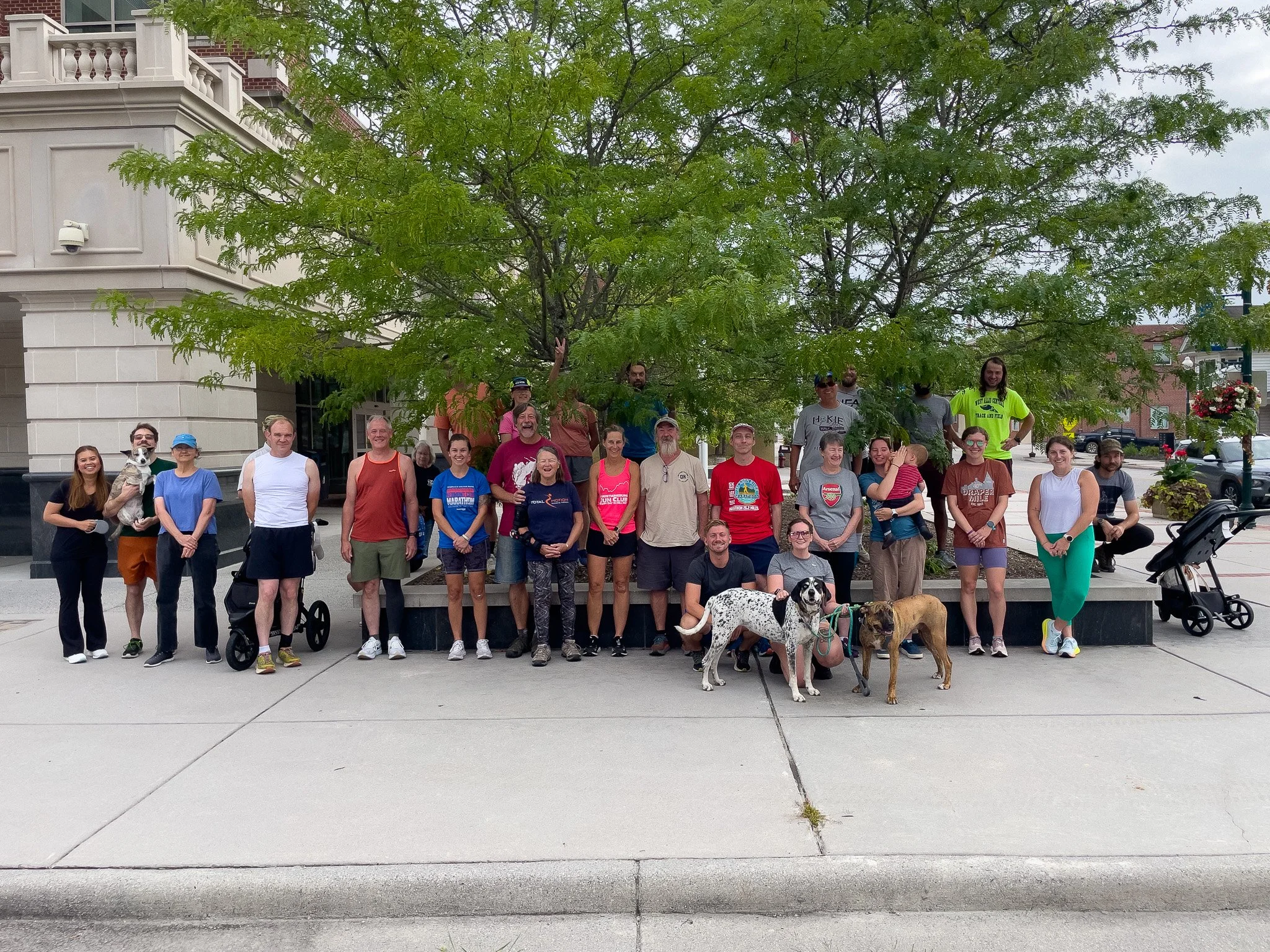 Group of people standing and sitting under a large tree on a city sidewalk, with some holding dogs, in front of a beige building.