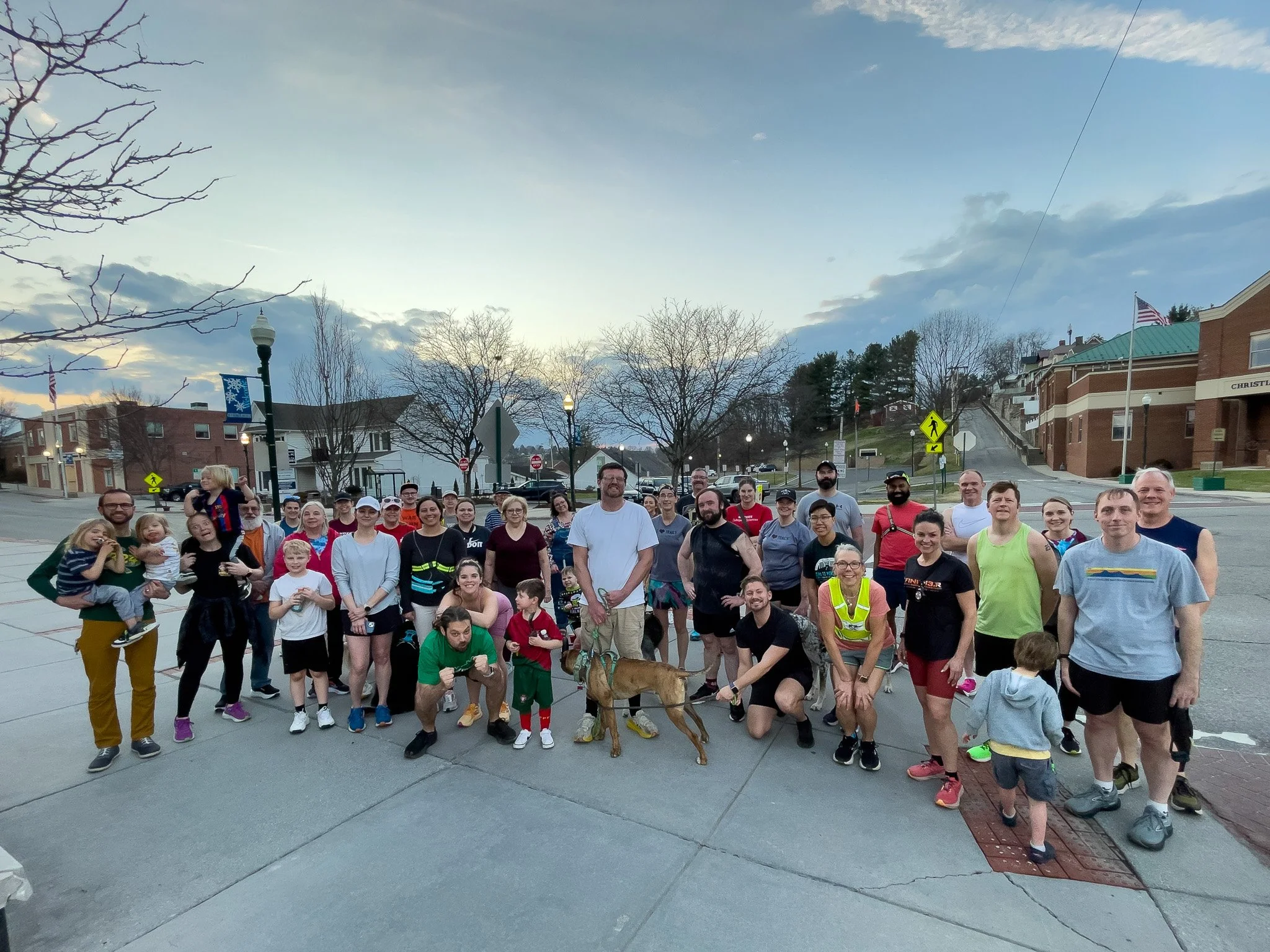 A large group of people gathered outdoors in a small town or neighborhood, with some children and a dog, during late evening or early morning, with trees, houses, and street signs in the background.