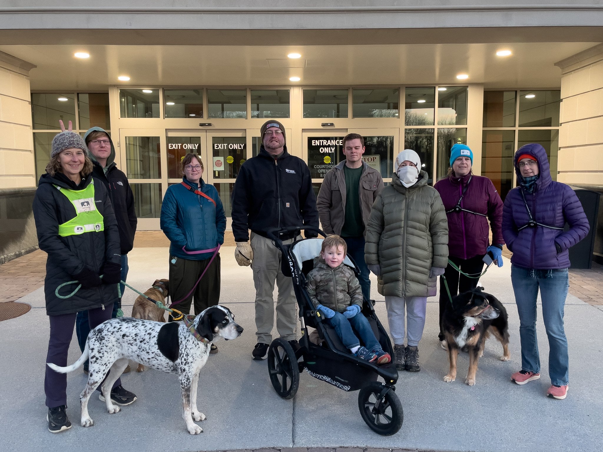 Group of nine people and three dogs standing outside of a building with glass doors labeled 'Exit Only' and 'Entrance Only.'