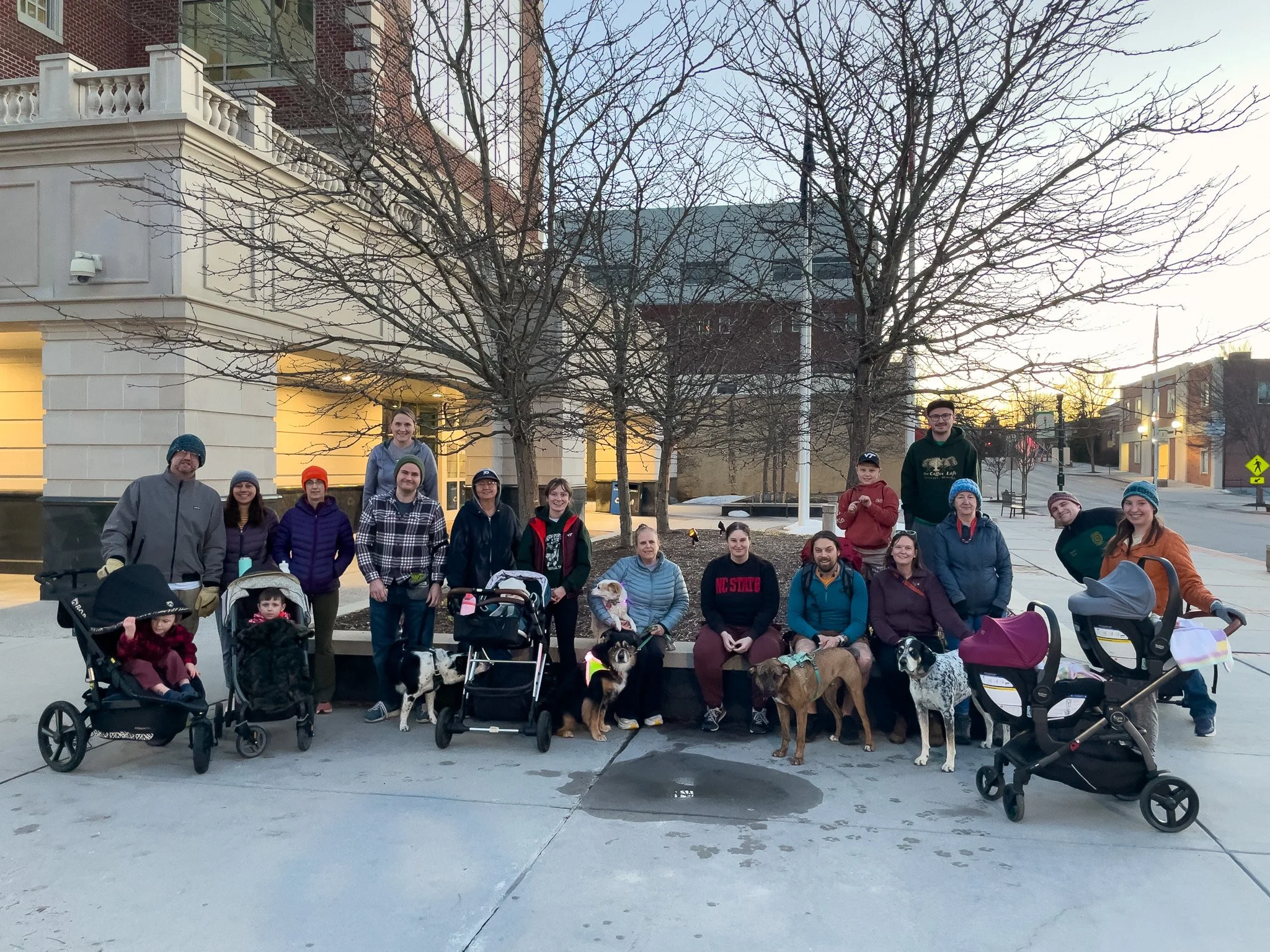 Group of people with dogs and strollers outdoors in front of a building and leafless trees during sunset.