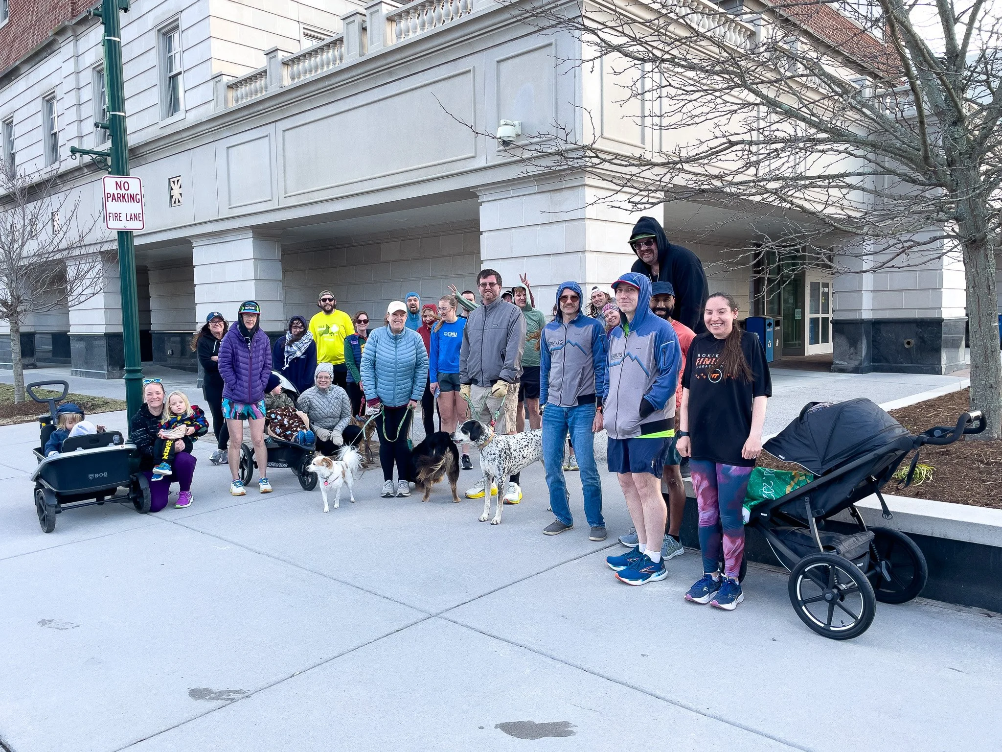 A group of people and dogs pose on a city sidewalk in front of a building during daytime. Some are wearing athletic clothing, and children are present. There are strollers and leashes, and a 