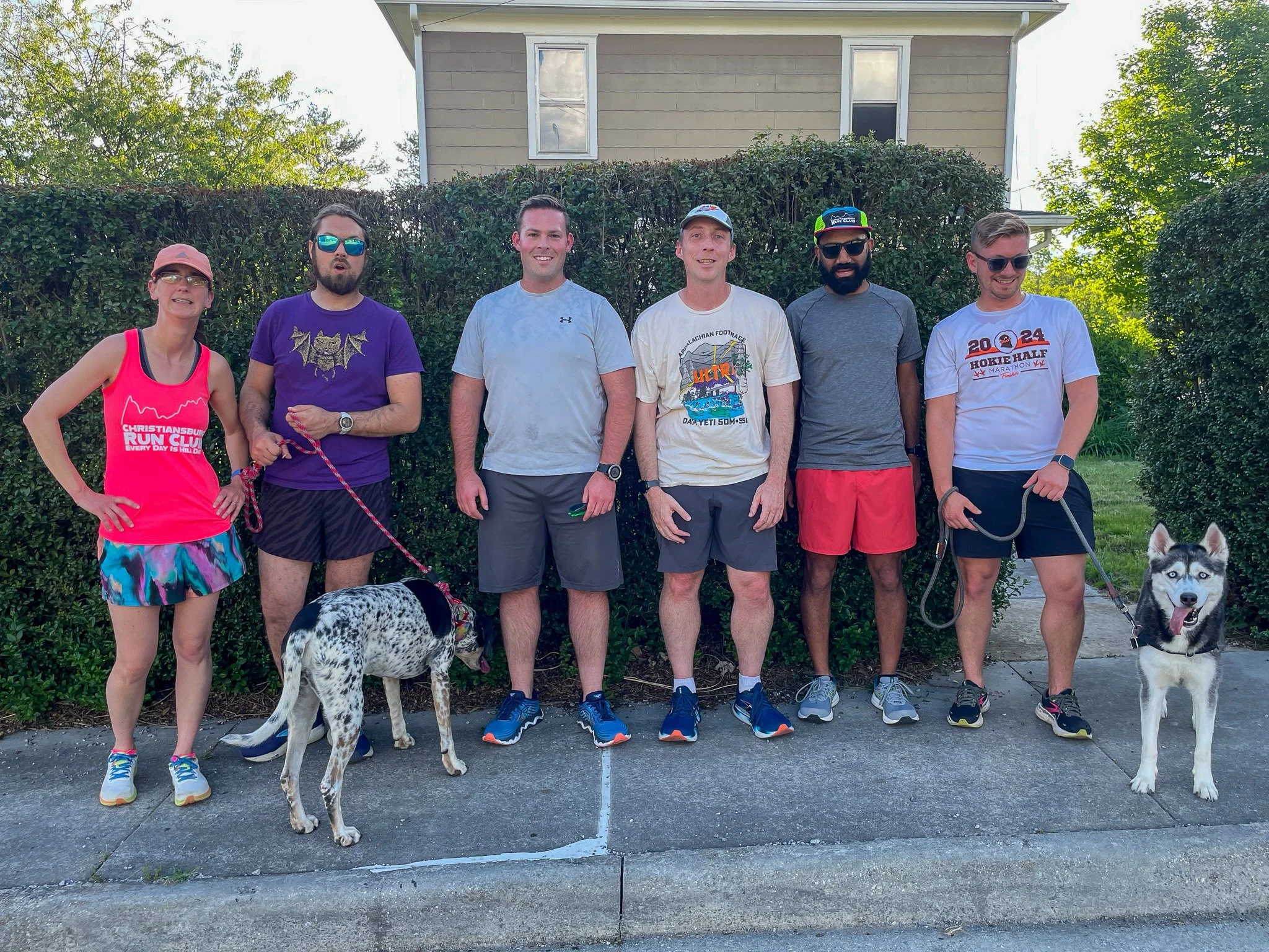 Group of six people standing outdoors with two dogs, three wearing sunglasses, in front of a hedge and a house.
