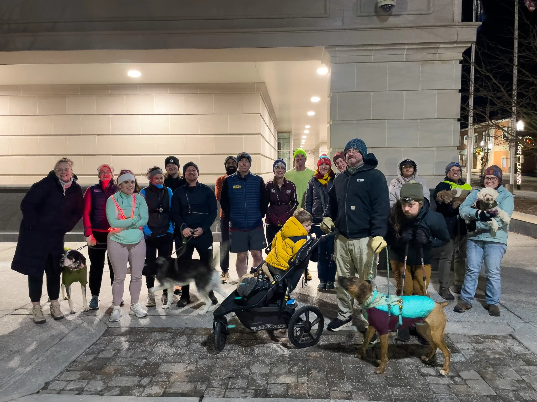 Group of people gathered outdoors at night, some with dogs, wearing winter clothing, standing on a paved area in front of a building.