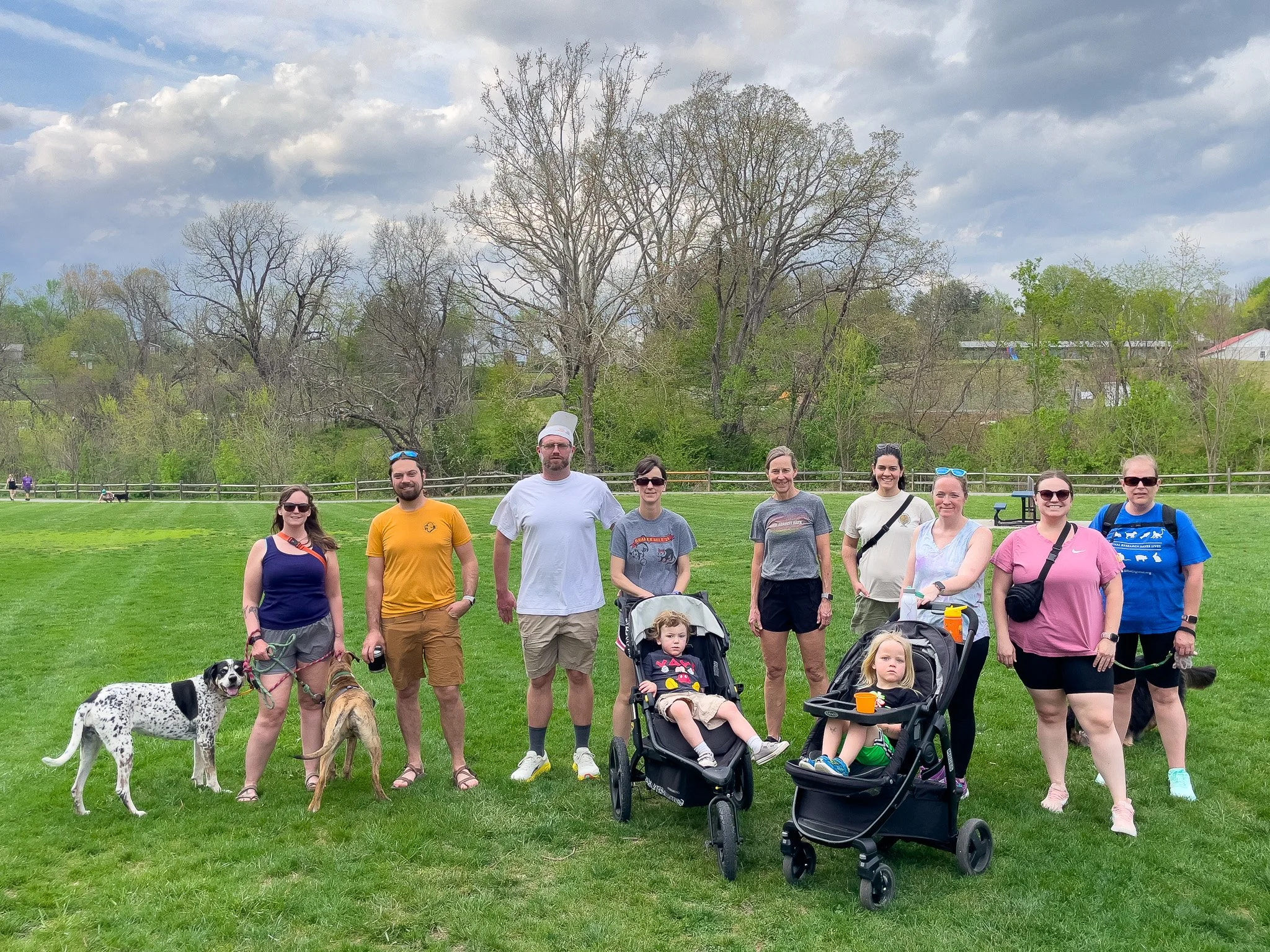 A group of ten people and four dogs standing on a grassy field with trees and a cloudy sky in the background.