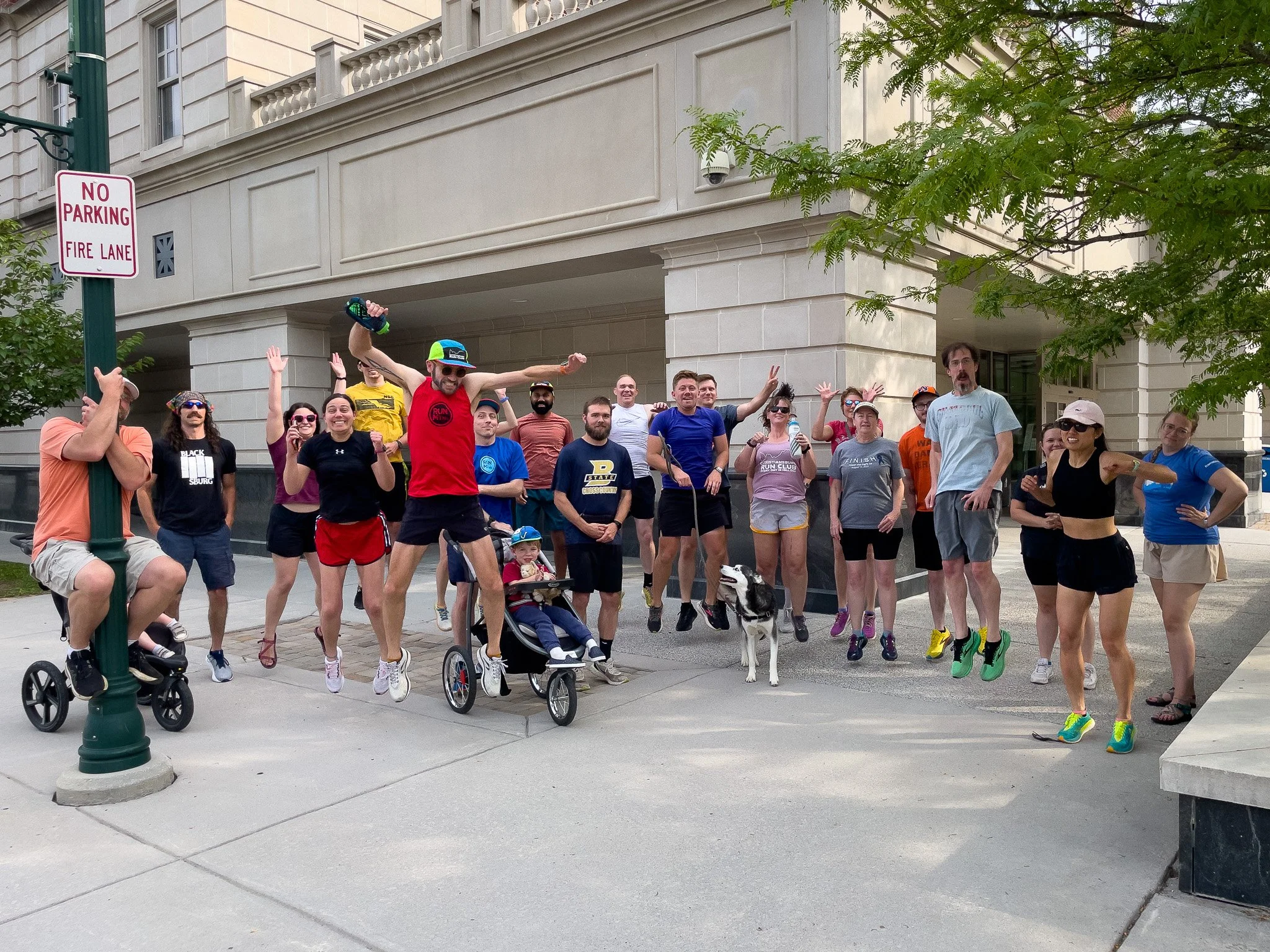 Group of people gathered outdoors in an urban area, some jumping and posing for a photo, with a man in a red tank top and a child in a stroller in front, alongside a Dalmatian dog, trees, and a beige building in the background.