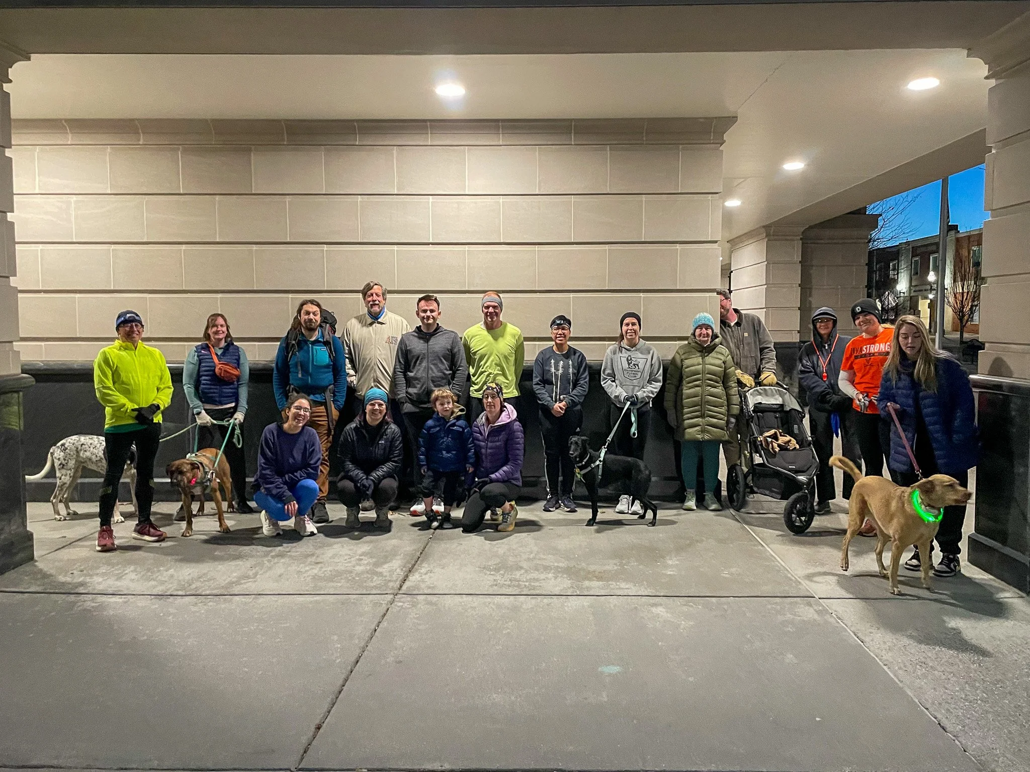 Group of people, some with dogs, gathered outdoors at night, standing and squatting in front of a large building wall under streetlights.