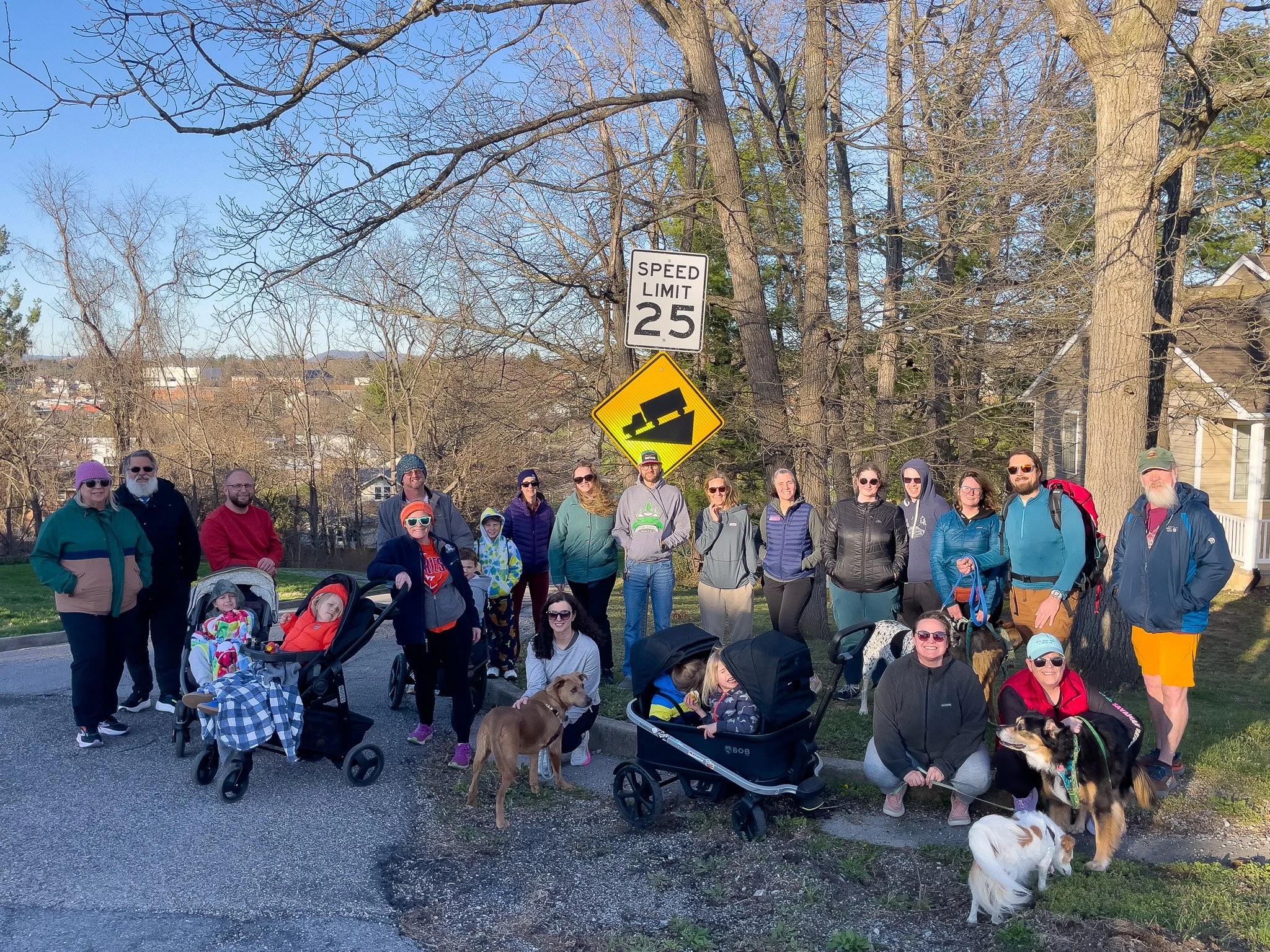 A group of people, including children and dogs, gathered outdoors on a street with leafless trees and a house in the background. The group is standing near a speed limit sign and a warning sign about a steep grade.