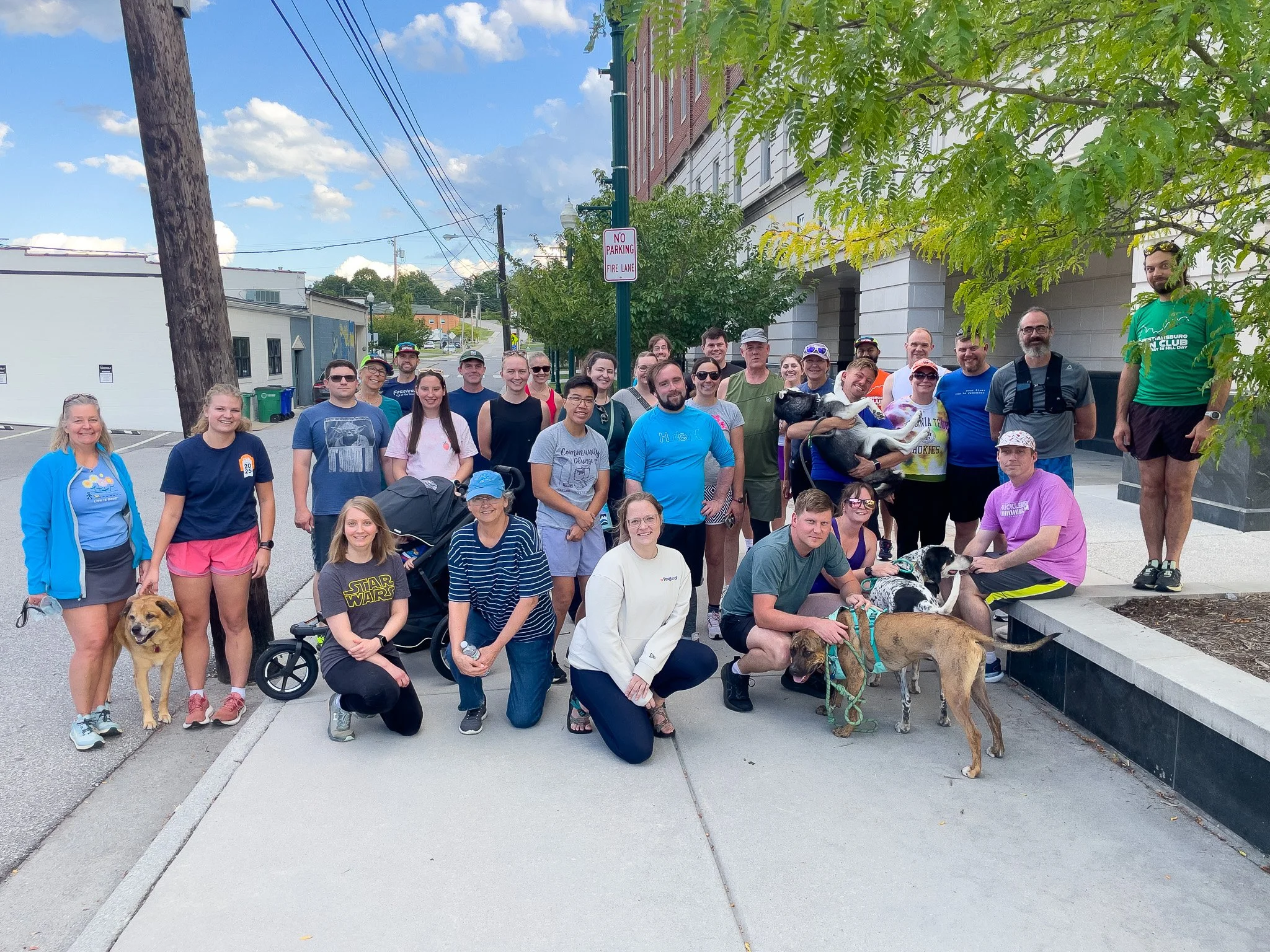 Group of people with dogs posing for a photo on a city sidewalk under a tree.
