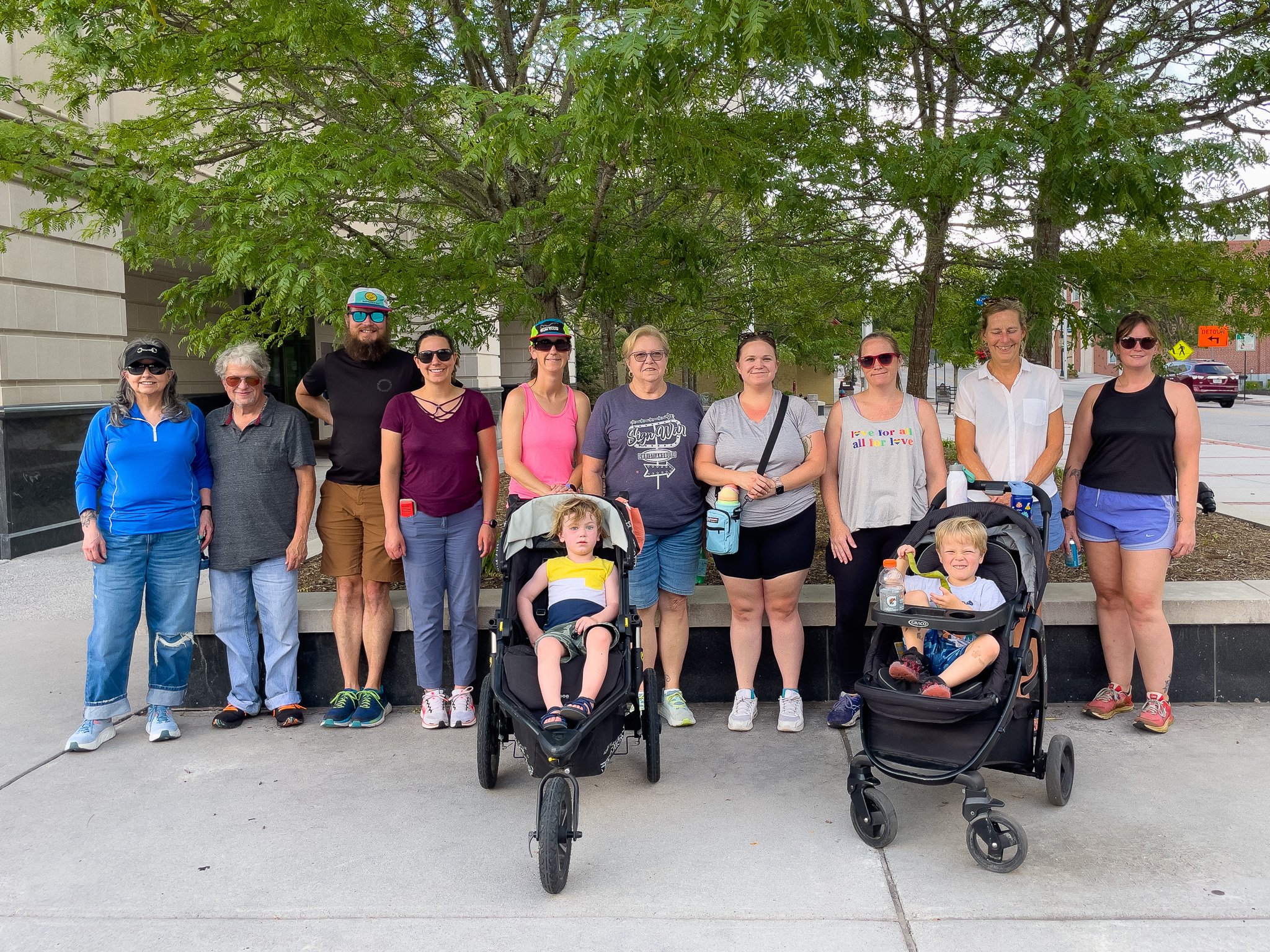 Group of 11 people, including children in strollers, standing outdoors in front of a tree and a building on a city sidewalk.