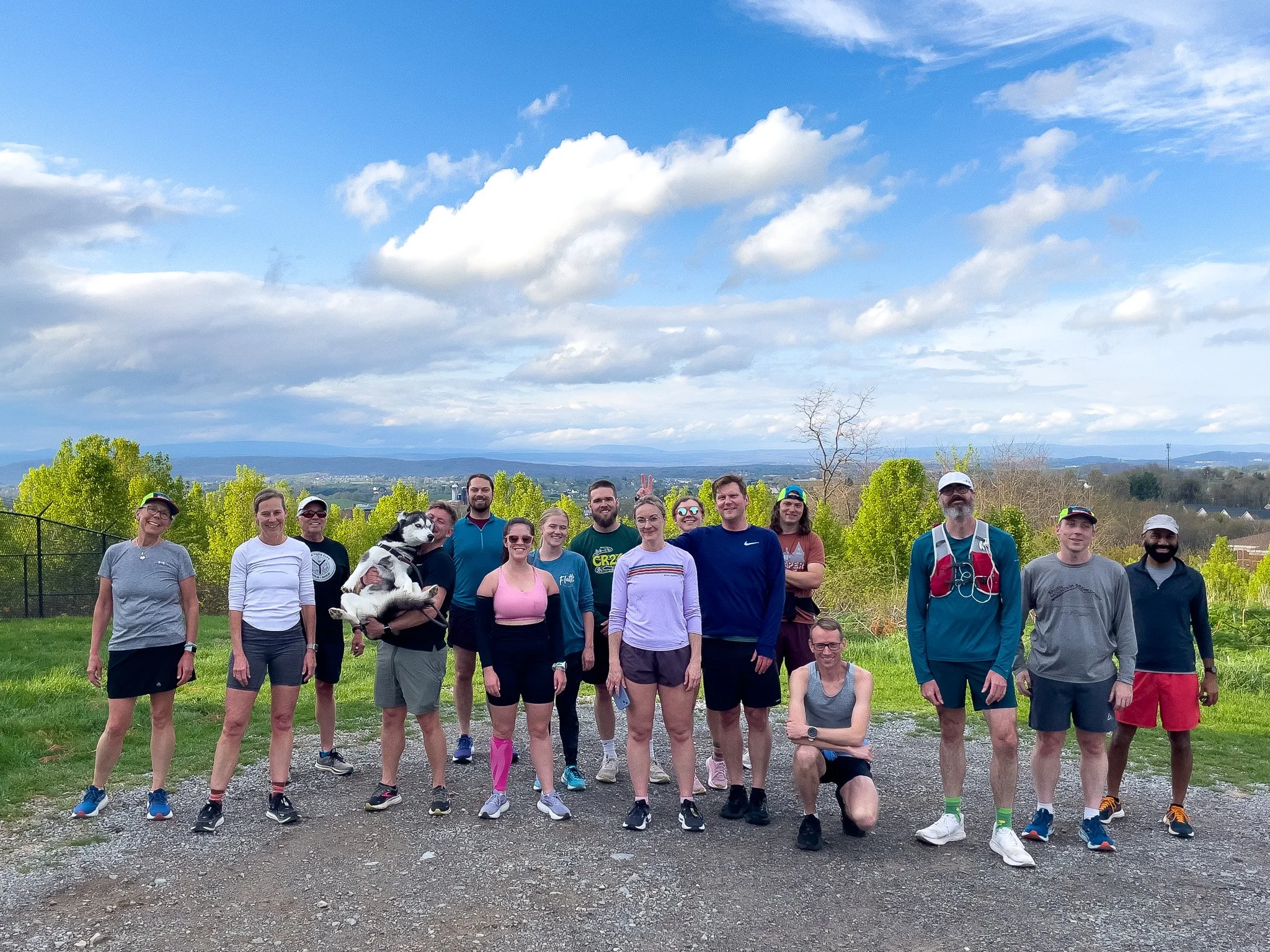 Group of 15 runners, including men and women of diverse ages, posing outdoors on a gravel path with greenery and mountains in the background. Some are smiling, one person is holding a husky dog, and one person is making a peace sign.