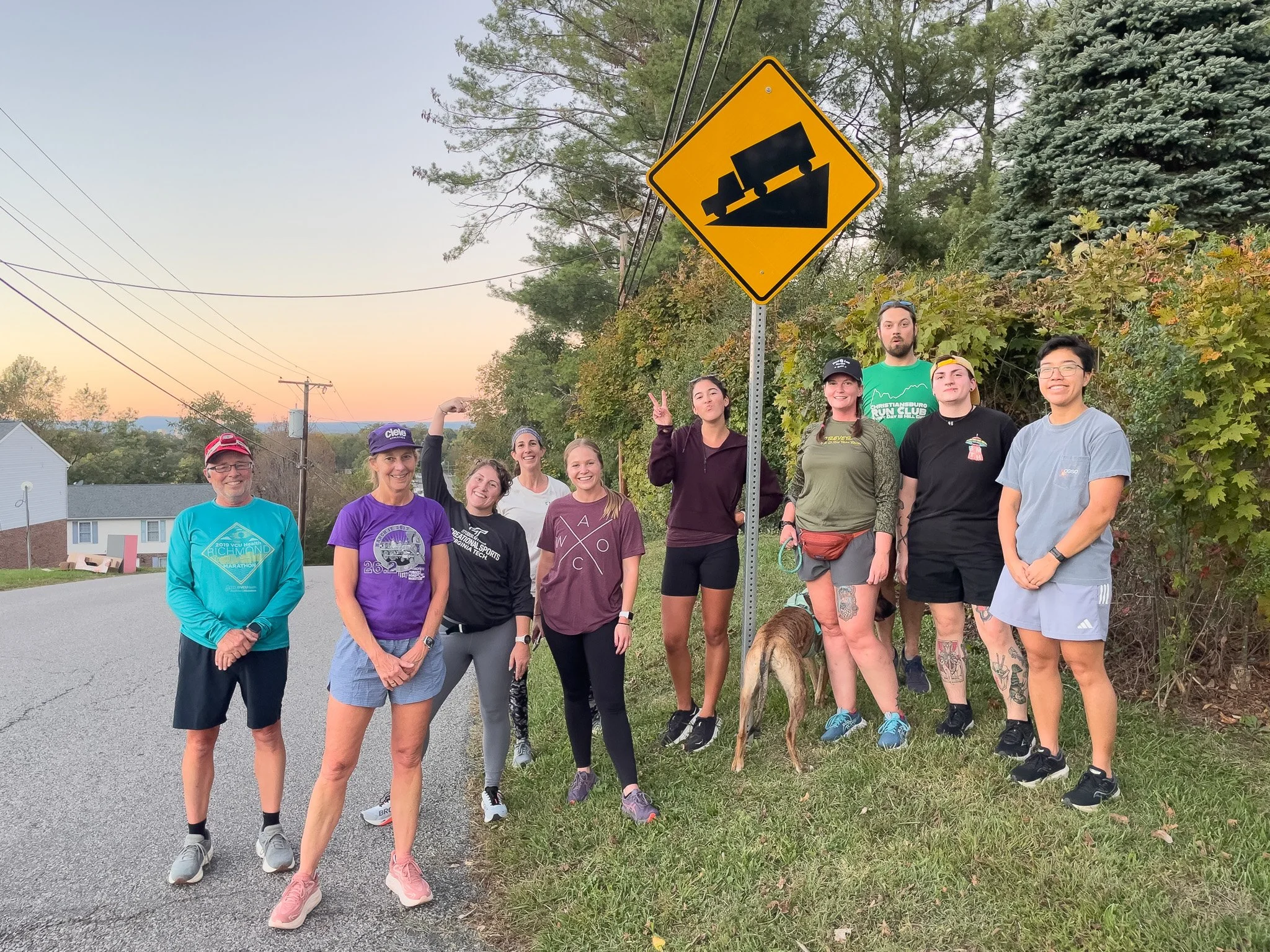 Group of nine people standing on the side of the road with a yellow road sign indicating a steep hill or incline and a train, possibly a railway crossing sign. They are outdoors during sunset, some smiling and making gestures.